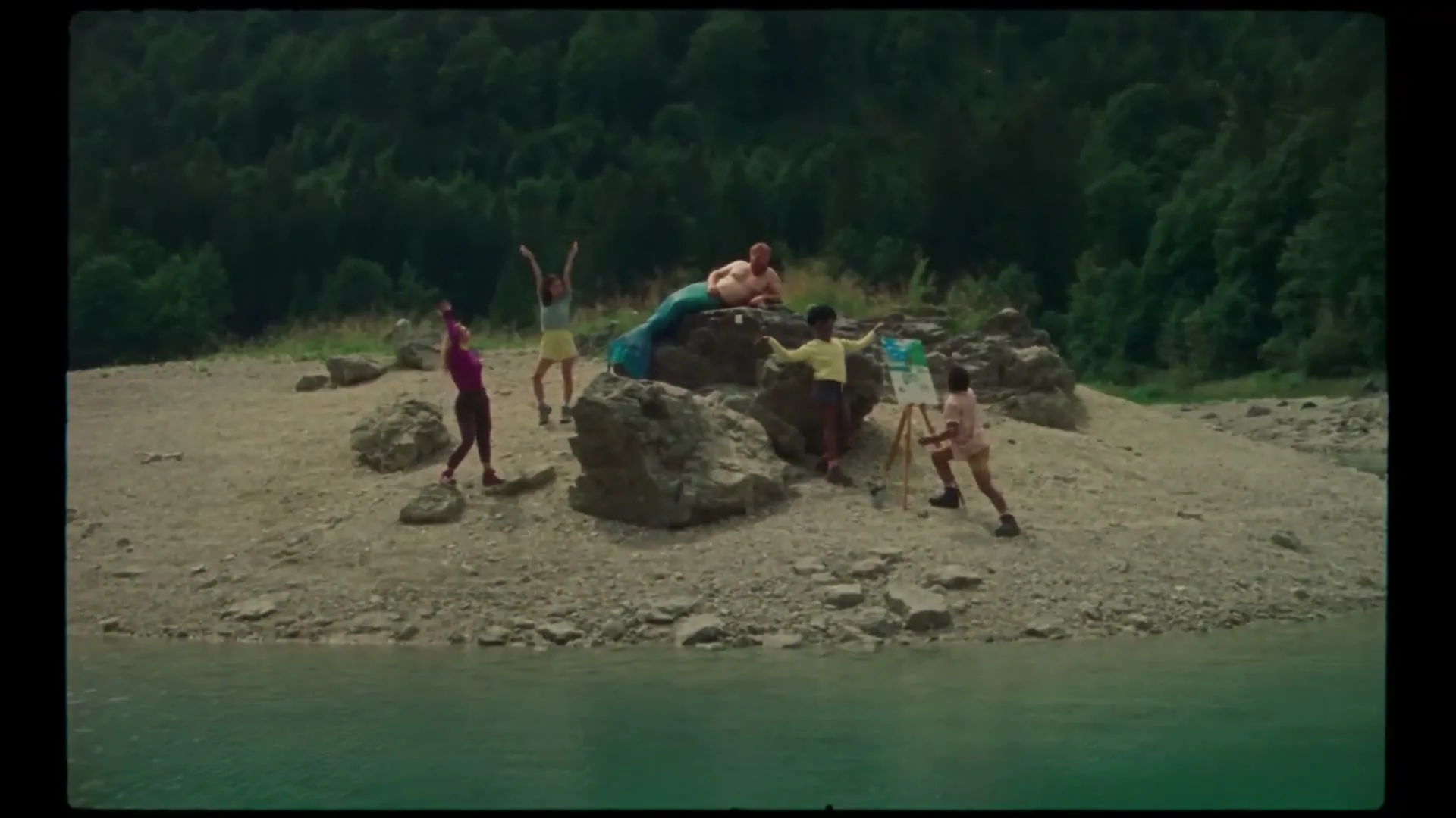 Five young adults pose and paint a person dressed as a mermaid on a rocky island in a lake, surrounded by green trees.