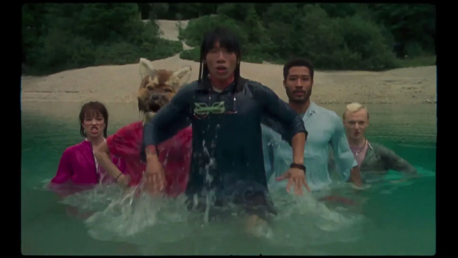 Four people and one person in a fox mask emerge from a river, splashing water, on a sunny day with a sandy bank and trees in the background.