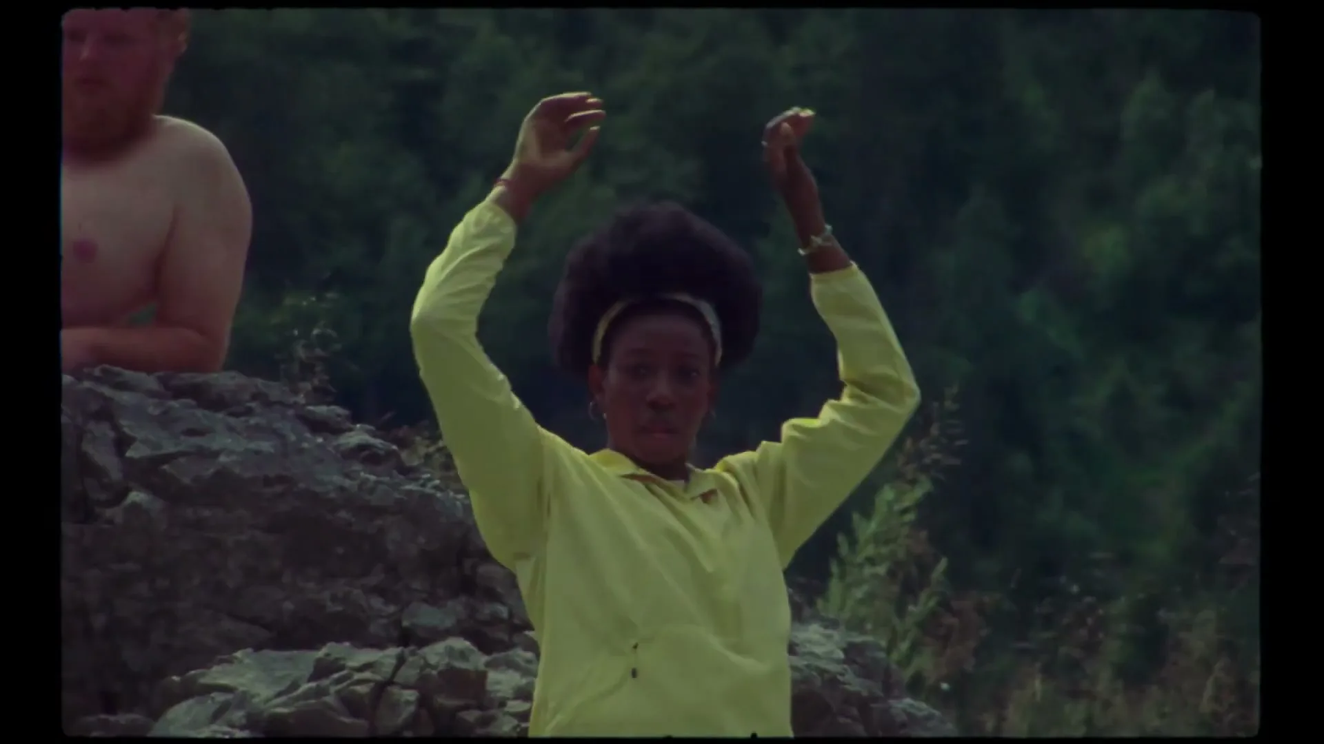 A woman in a yellow jacket stretches her arms upwards, while a man sits shirtless in the background on a rocky, forested hillside.