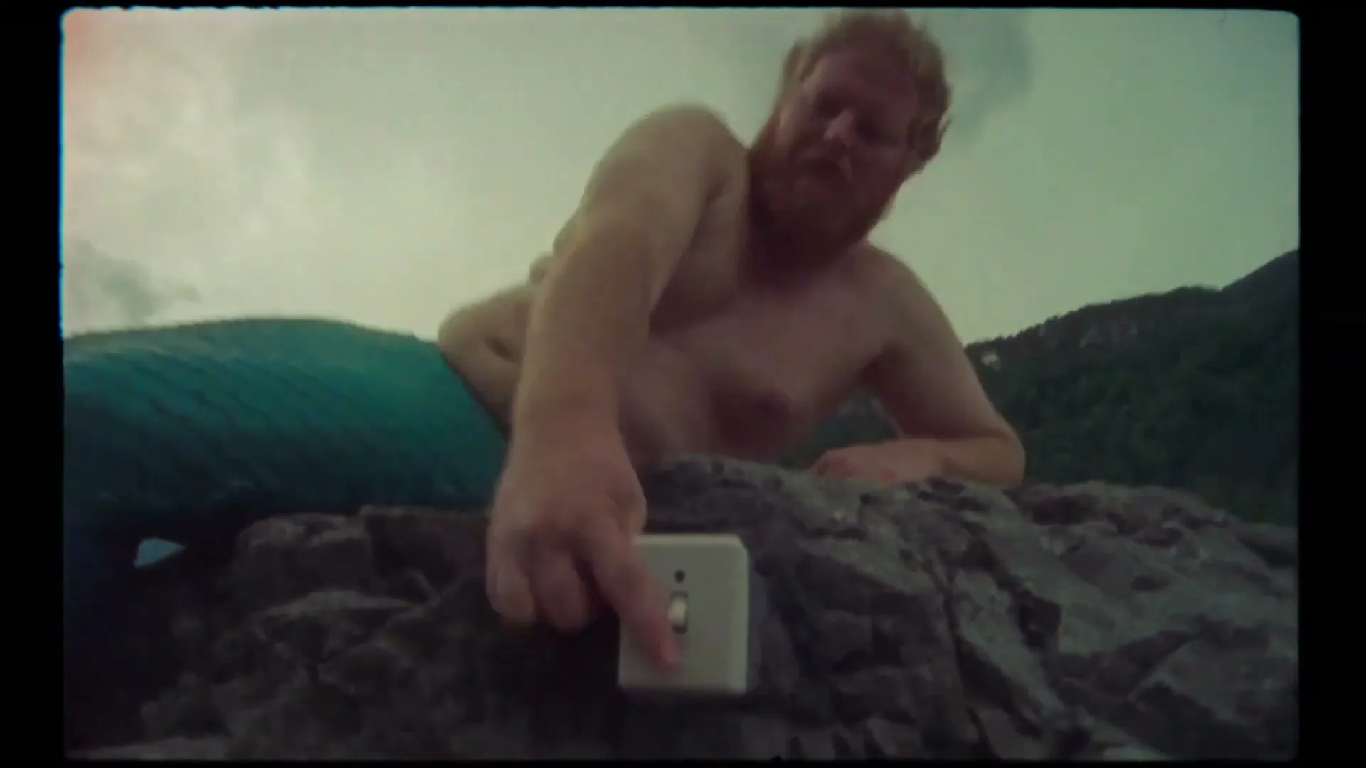 A shirtless, bearded man with a mermaid tail lies on rocks, reaching to flip a light switch under a cloudy sky.