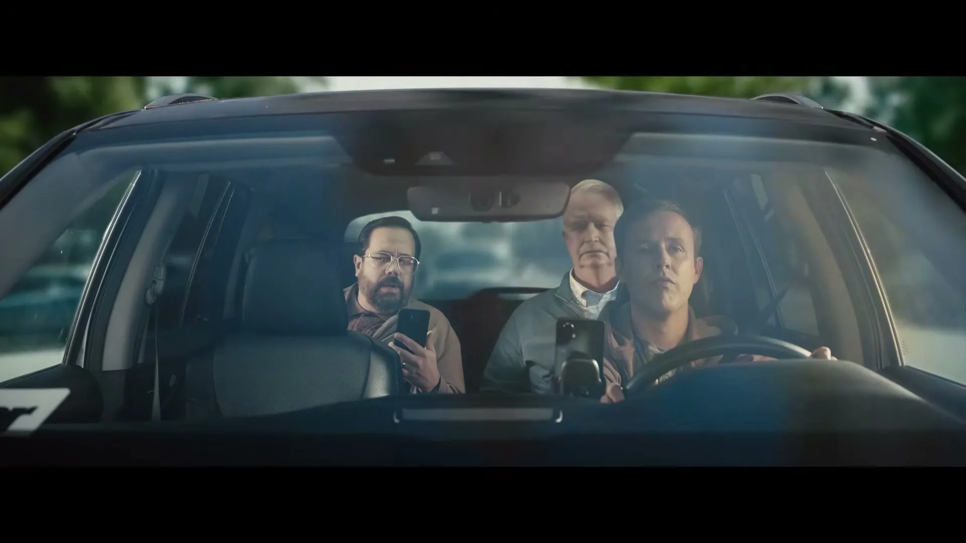 Three men are seen through the windshield of a car, with the driver looking ahead and the two passengers looking at their phones.