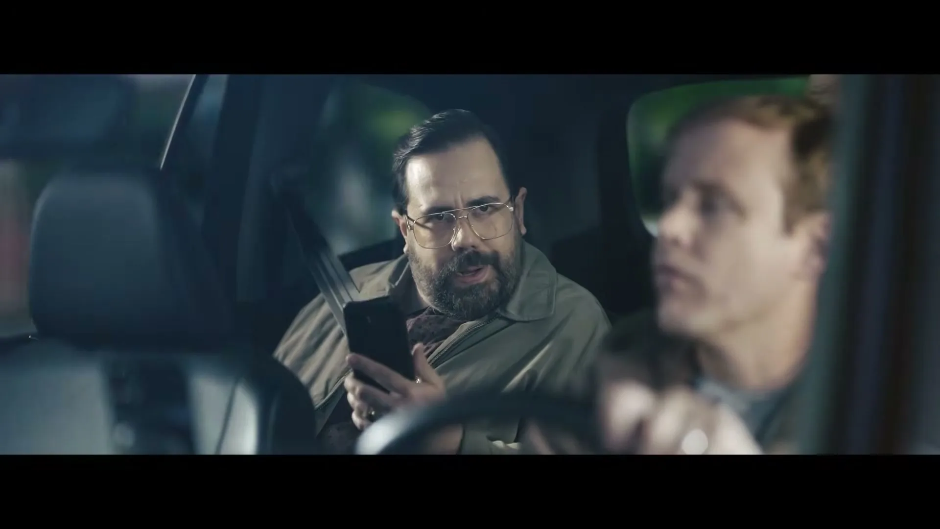 A man with a beard and glasses looks at his phone in the passenger seat of a car, while another man drives.