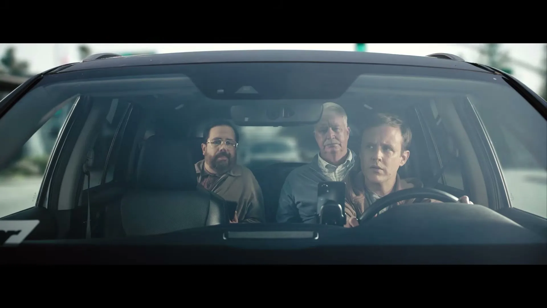 Three men, one driving, one in the passenger seat, and one in the back, are seen through the windshield of a car, driving on a sunny day.