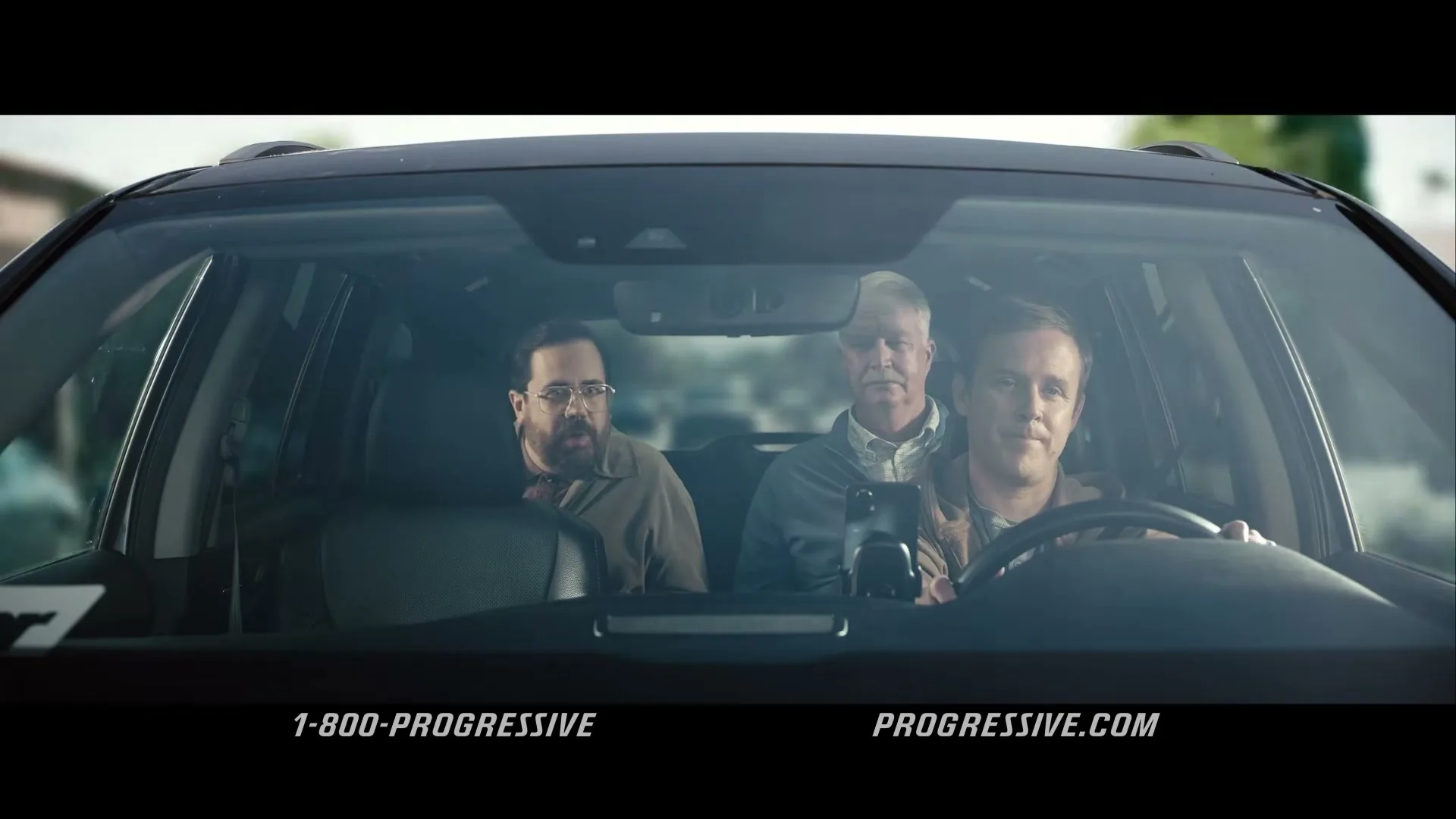 Three men are seen through the windshield of a car, with the driver looking forward and the two passengers looking at each other, as they drive down a road.