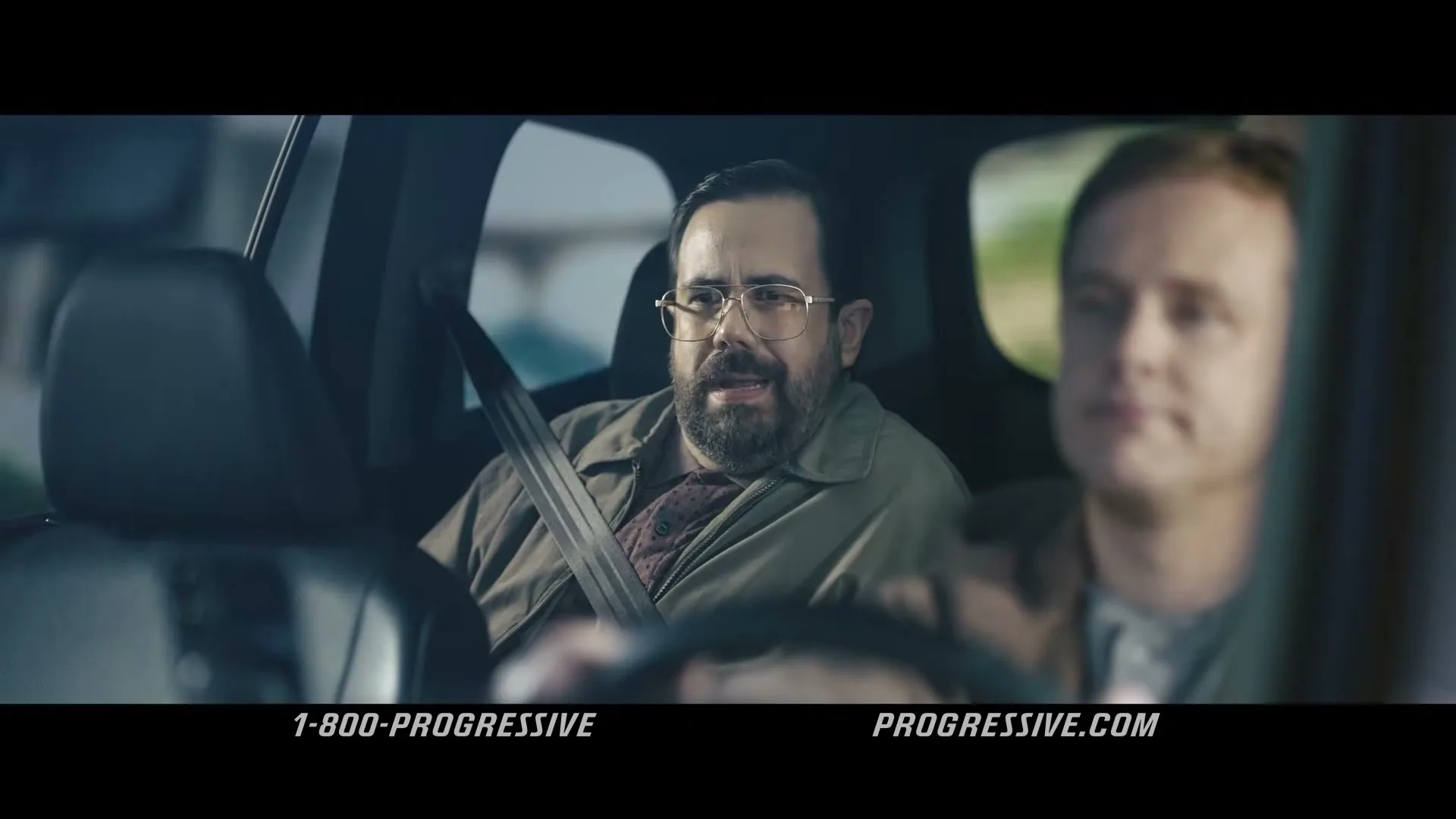A man with a beard and glasses sits in the back seat of a car, looking at the driver, who is out of focus.