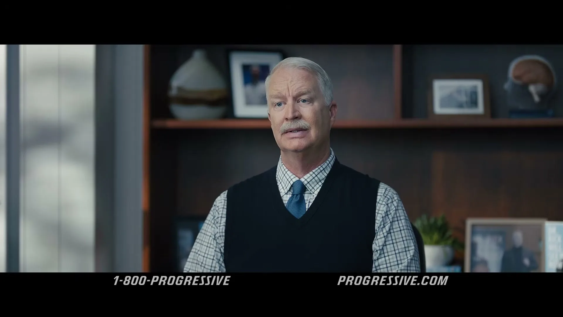 A middle-aged man with a mustache, wearing a sweater vest and tie, speaks directly to the camera in an office setting.