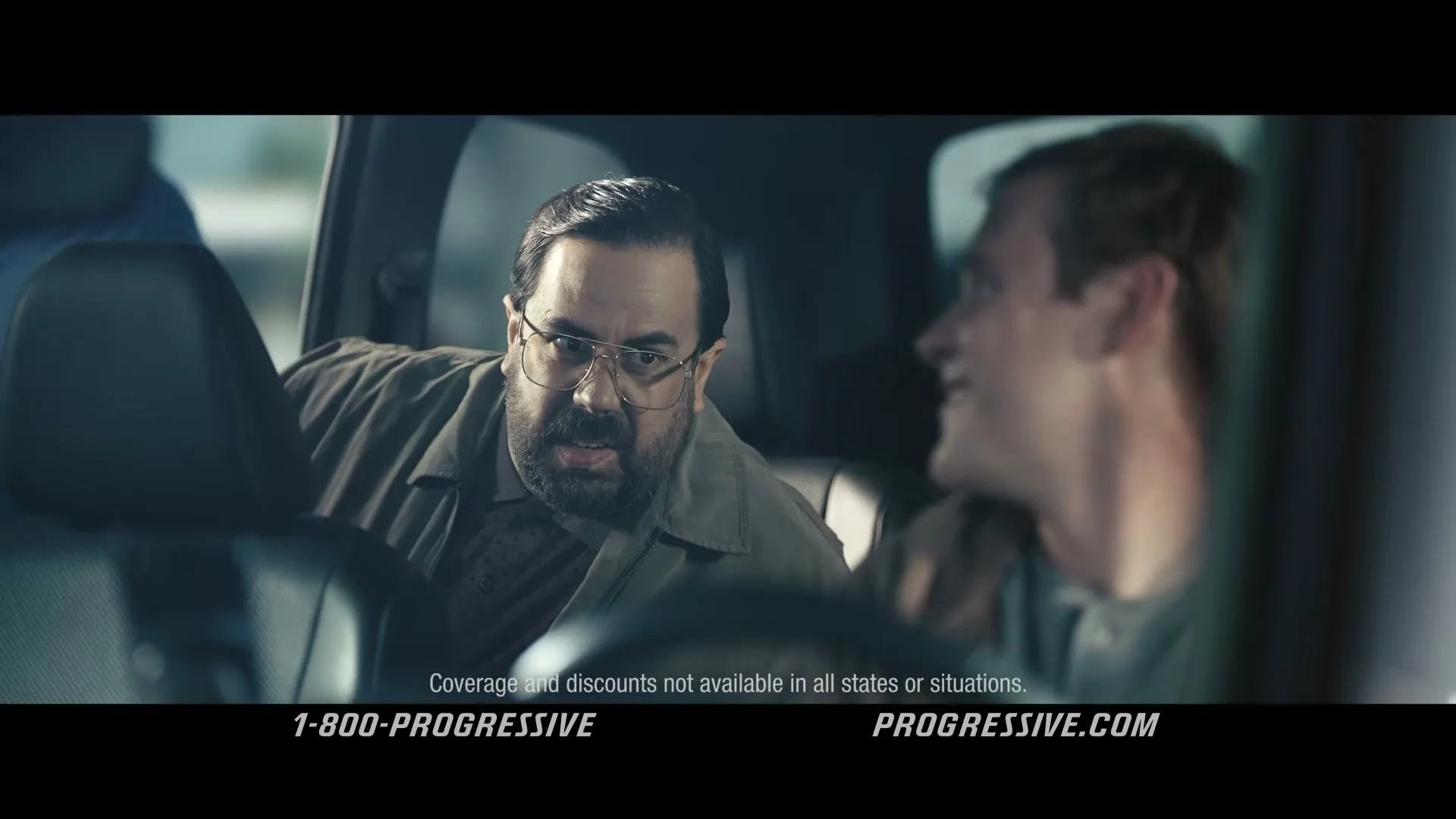 A man with a beard and glasses in a brown jacket looks at another man in the passenger seat of a car, who is smiling.