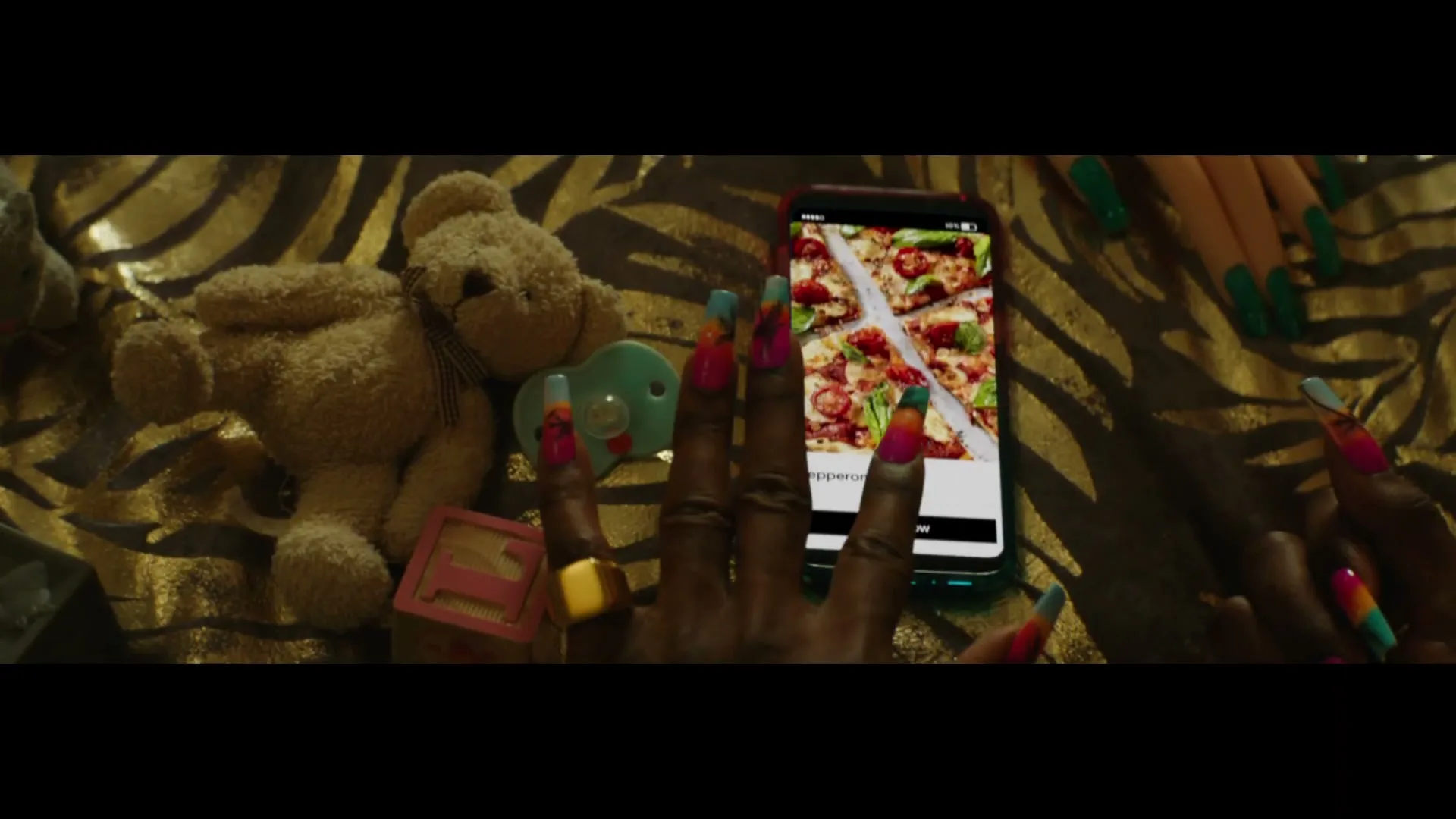 A woman with long, colorful nails scrolls through the Uber Eats app on her phone, surrounded by a teddy bear and baby pacifier on a tiger-print blanket.