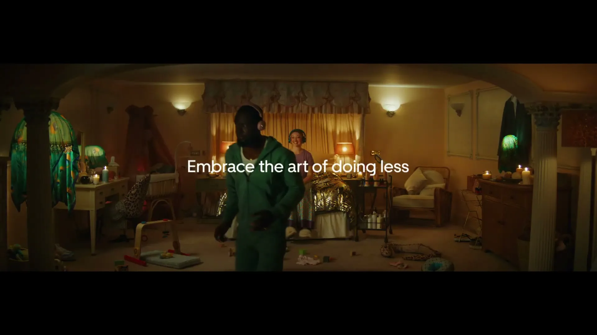 A man in a green tracksuit walks across a messy bedroom while a woman sits on the bed, both wearing headphones, with text on screen.