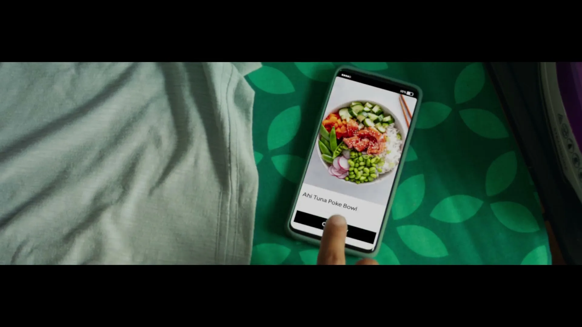 A person's hand taps on a smartphone screen, ordering an Ahi Tuna Poke Bowl from the Uber Eats app, on a green patterned surface.