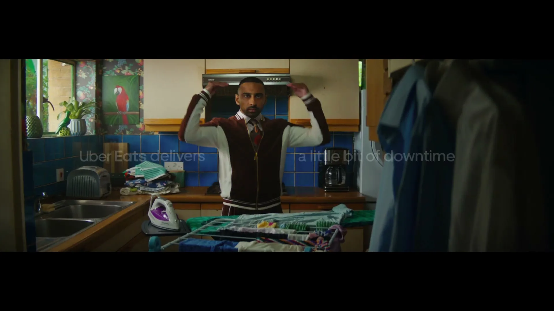 A man in a varsity jacket stands in a kitchen, looking at a pile of laundry on an ironing board, then puts on a white shirt.