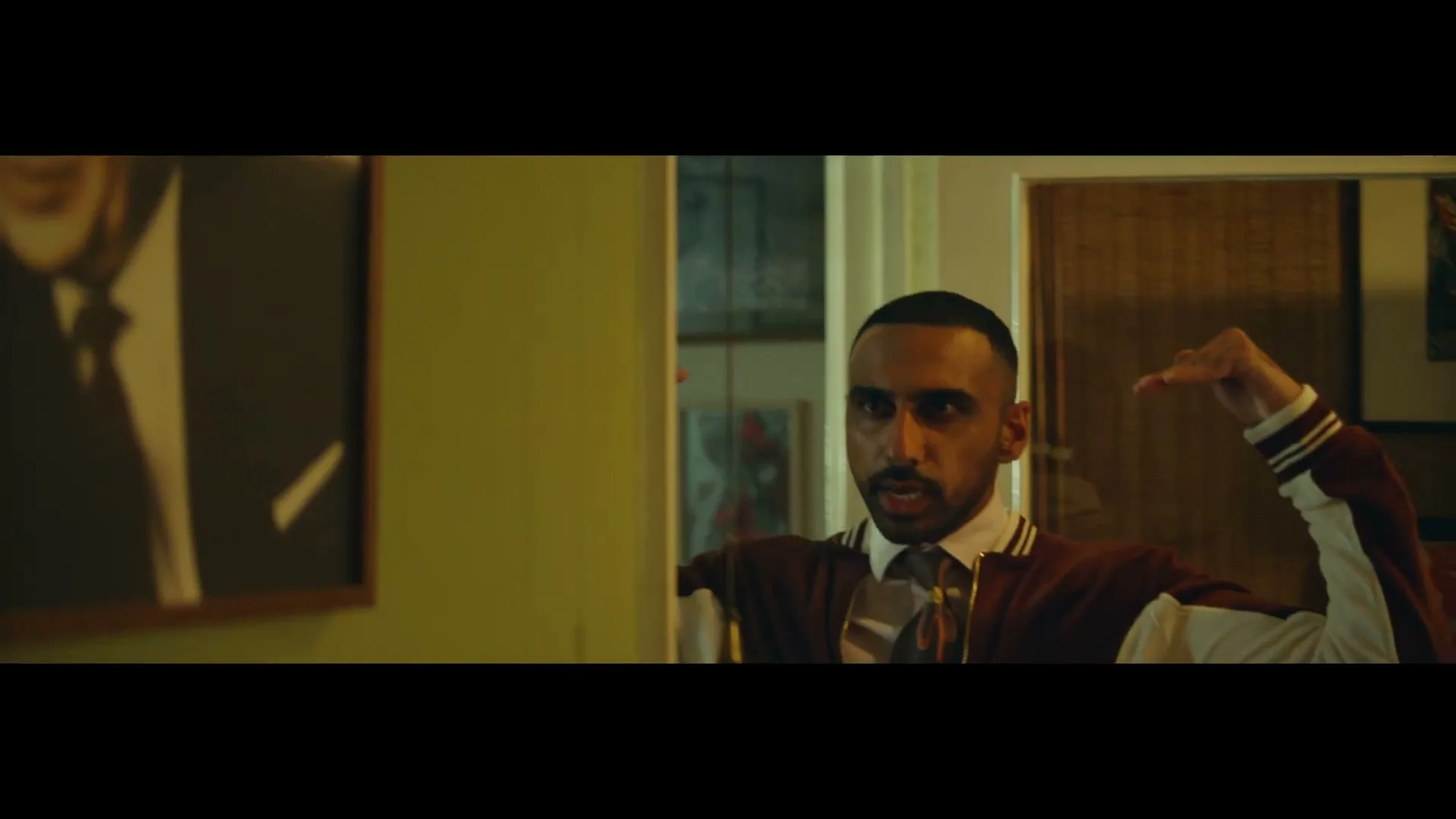 A man in a varsity jacket and tie stands in a room, making hand gestures as if mimicking a bird, next to a framed portrait on the wall.