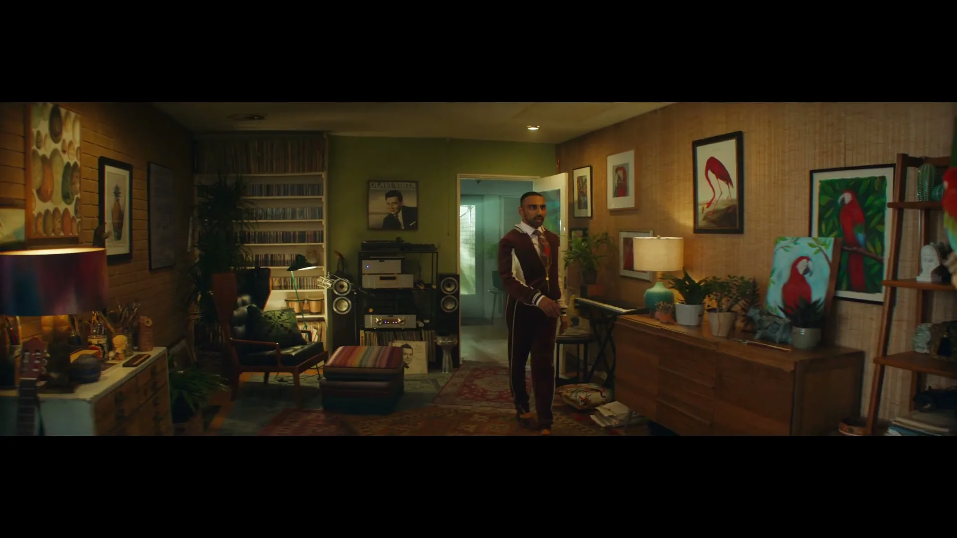 A man in a retro tracksuit sings karaoke in his living room, while a large parrot watches from a perch.