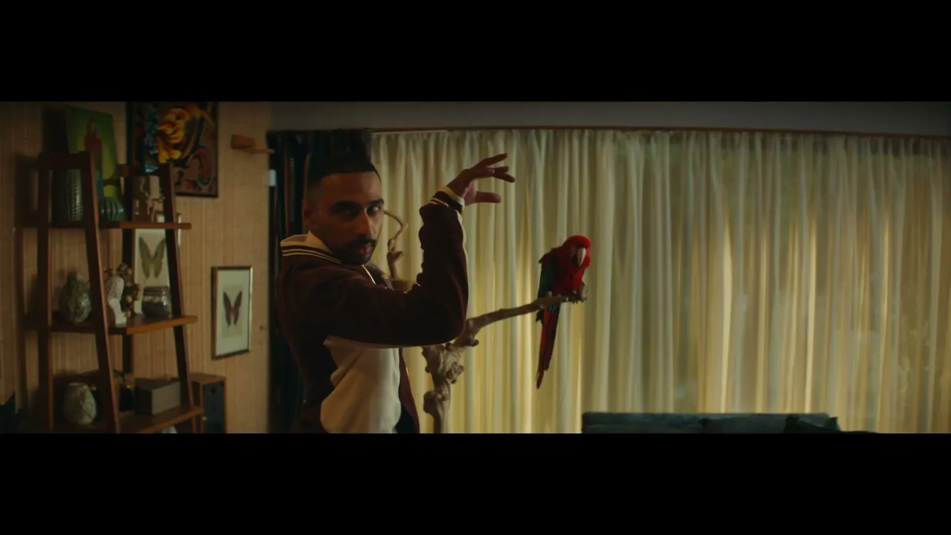 A man in a maroon and white sweater stands next to a red parrot on a perch, gesturing with his hands in a dimly lit room.
