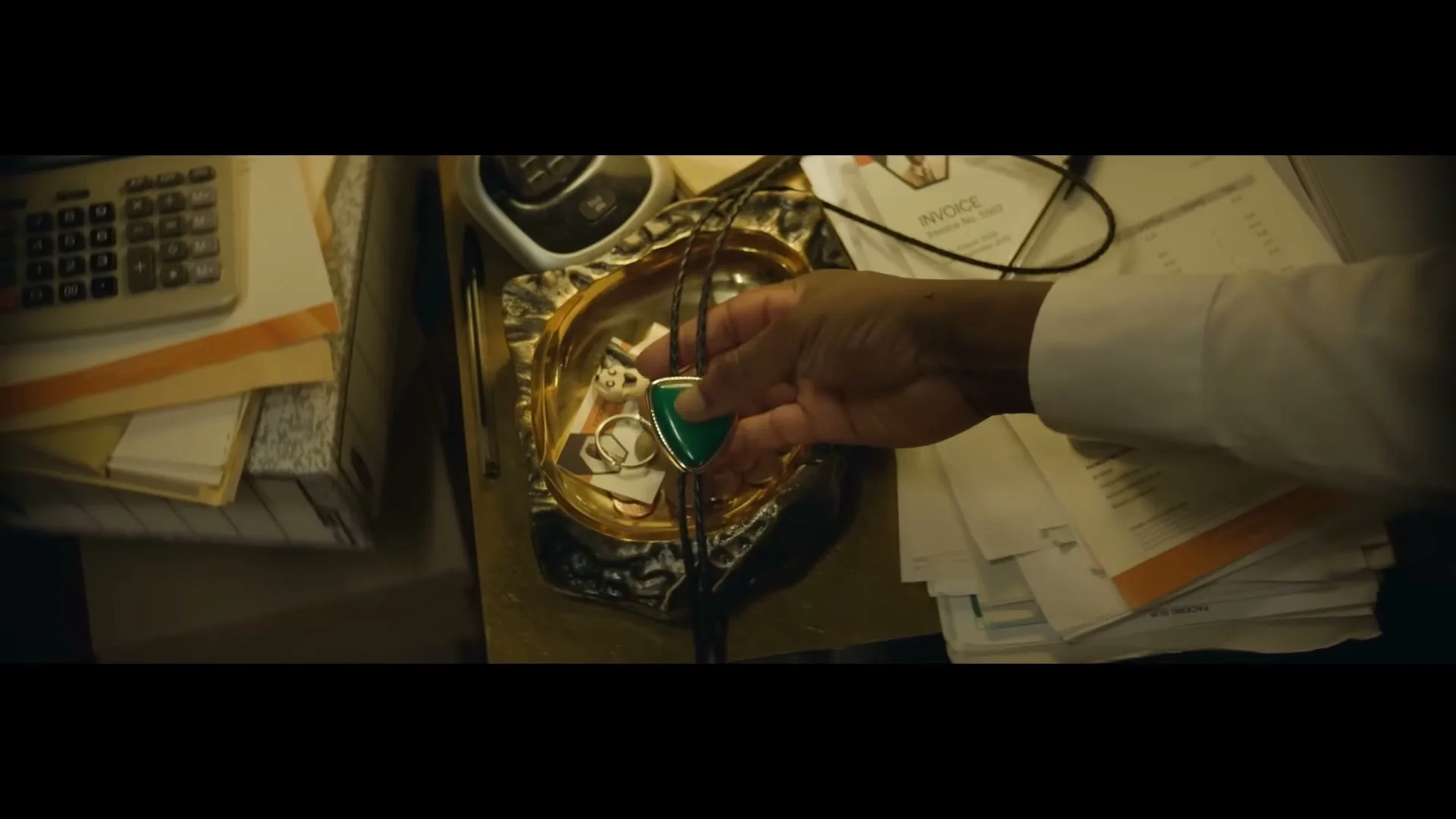 A man's hand picks up a bolo tie with a green stone from a decorative dish filled with coins and other small items on a cluttered desk.