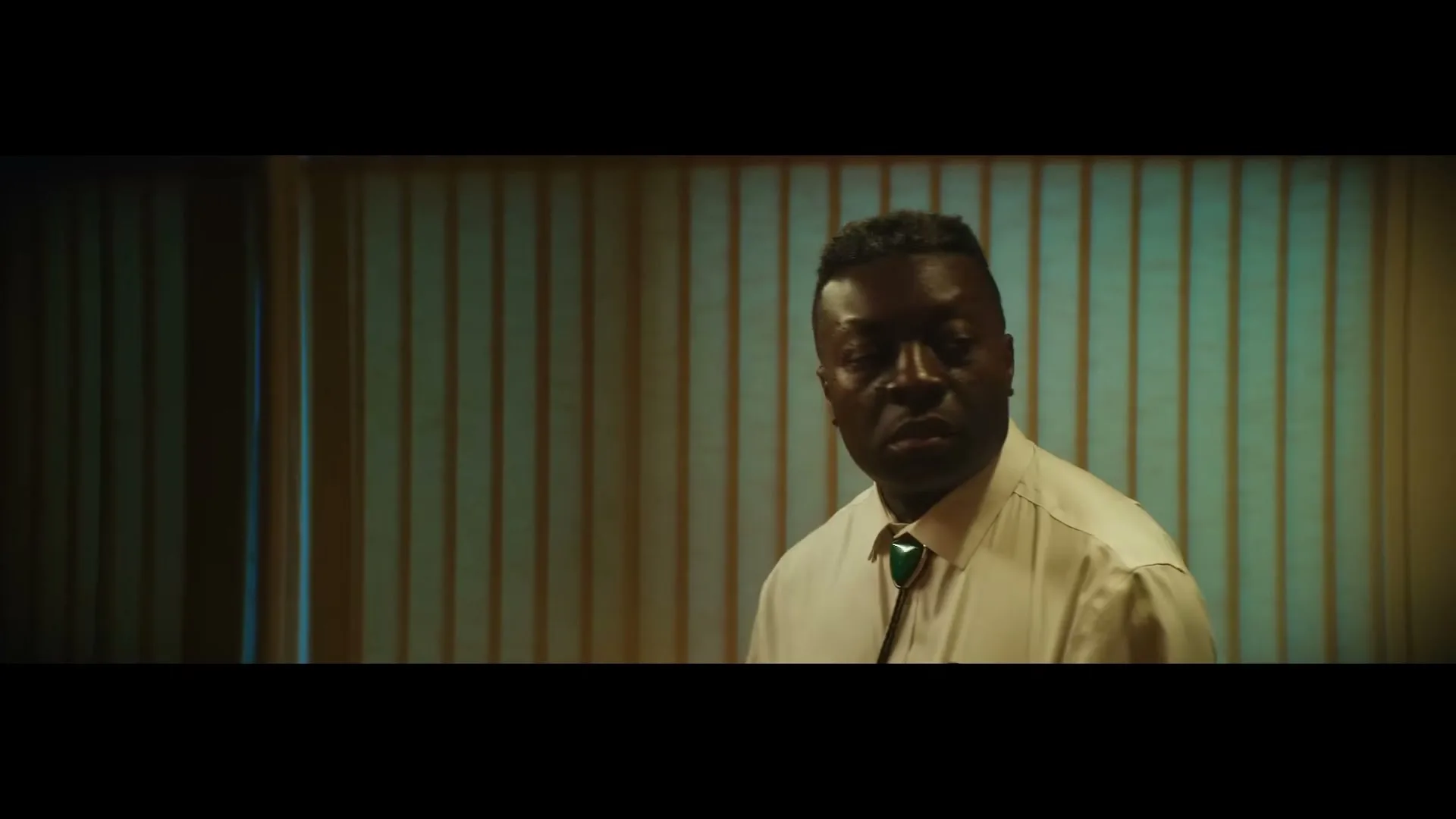 A Black man in a white shirt and bolo tie looks around a room with vertical blinds, adjusting his tie.