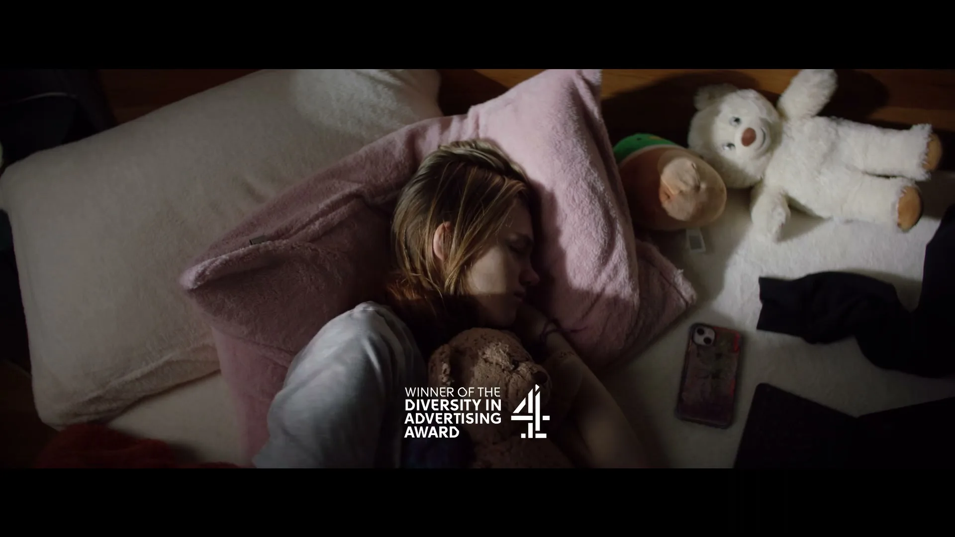 A young woman with blonde hair sleeps soundly in her bed, clutching a teddy bear, surrounded by pillows and other stuffed animals.