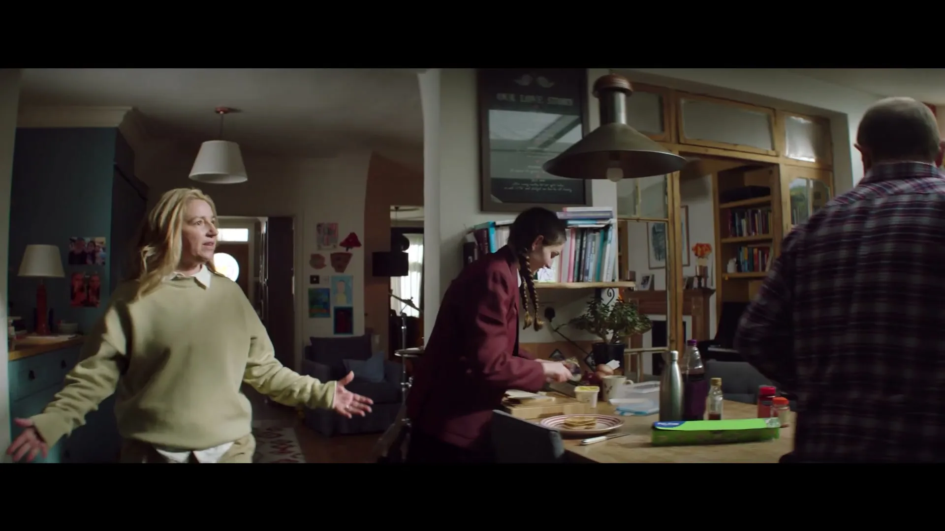 A family of three is in a kitchen/dining area, with a woman in a green sweater gesturing, a teen girl making food, and a man in a plaid shirt walking away.