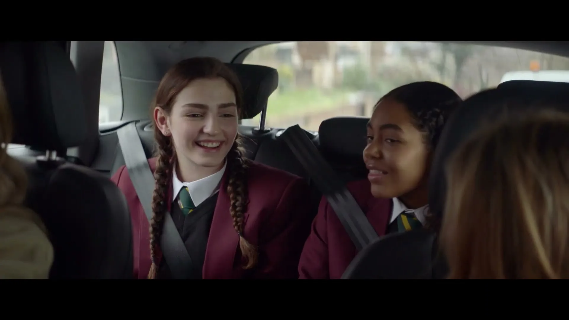 Two teen girls in school uniforms sit in the back of a car, talking and laughing, as seen from the front passenger seat.