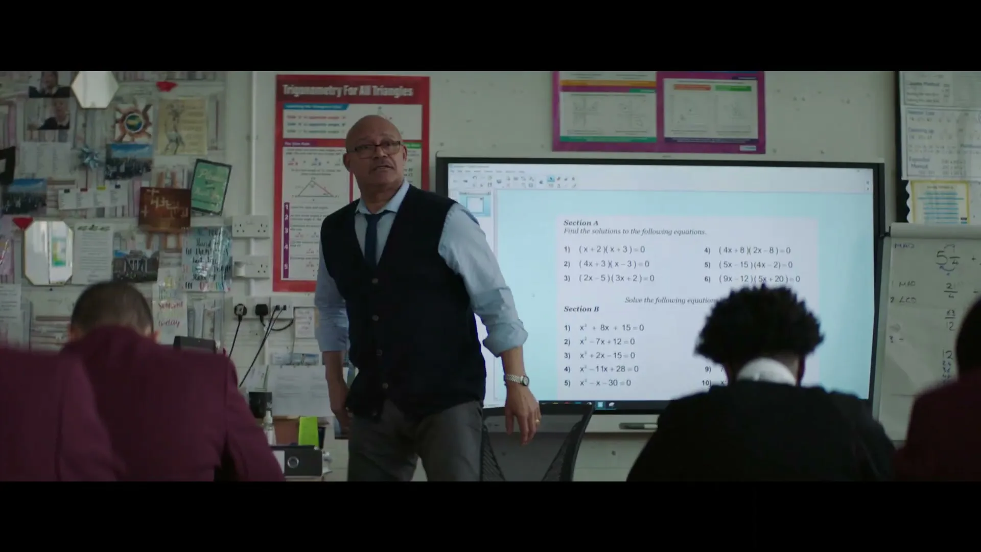 A male teacher in a blue shirt and dark vest walks around a classroom, looking at students while a math problem is displayed on a large screen.