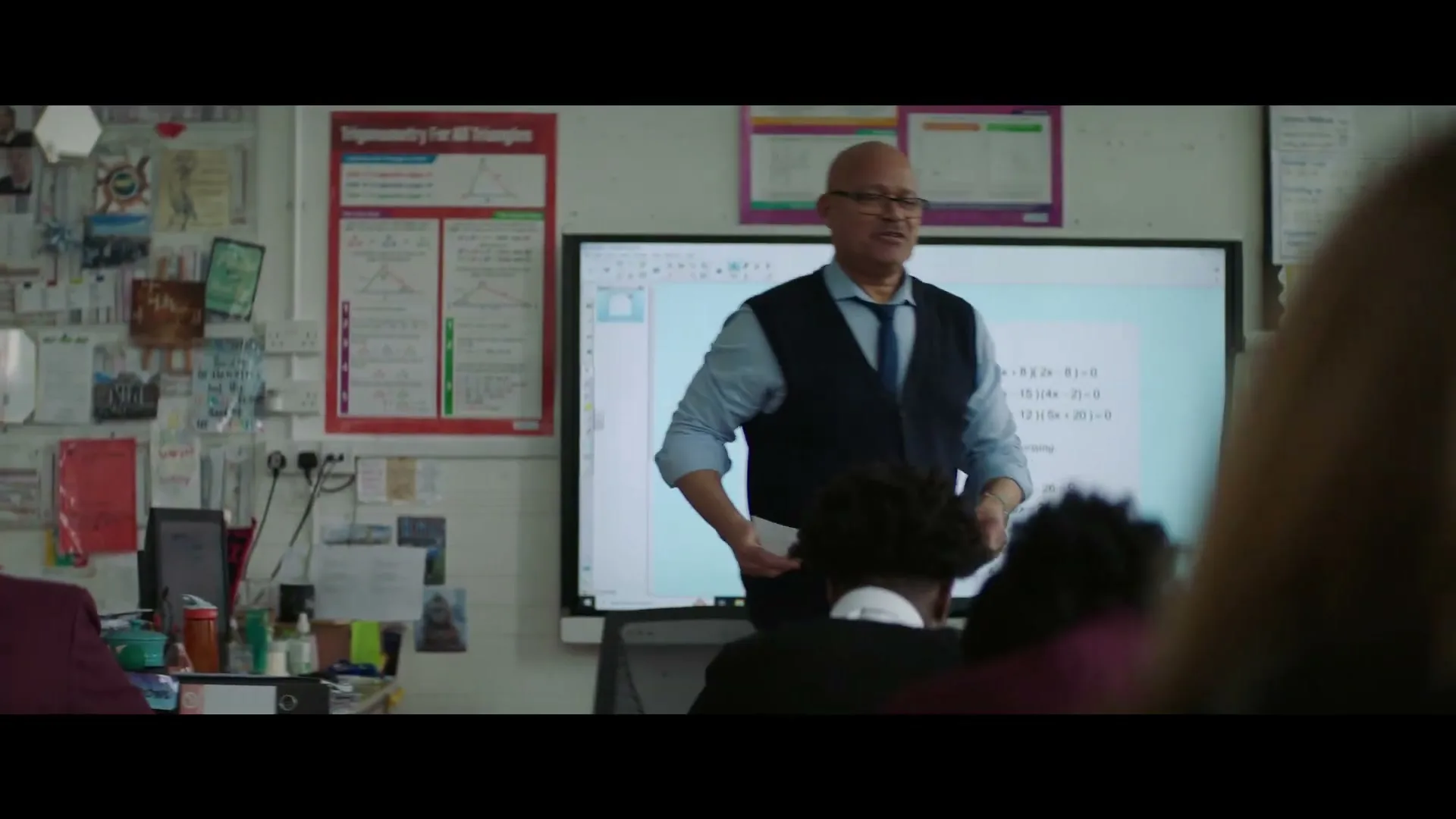 A male teacher, wearing glasses and a vest, stands in front of a whiteboard displaying math problems, addressing students in a classroom.