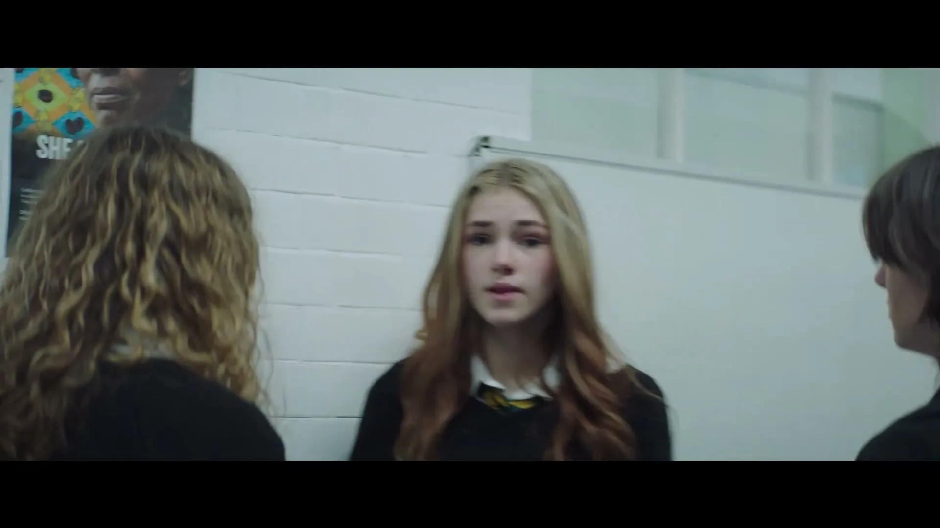 Three teens in school uniforms stand in a hallway, two with their backs to the camera, one facing them with a worried expression.