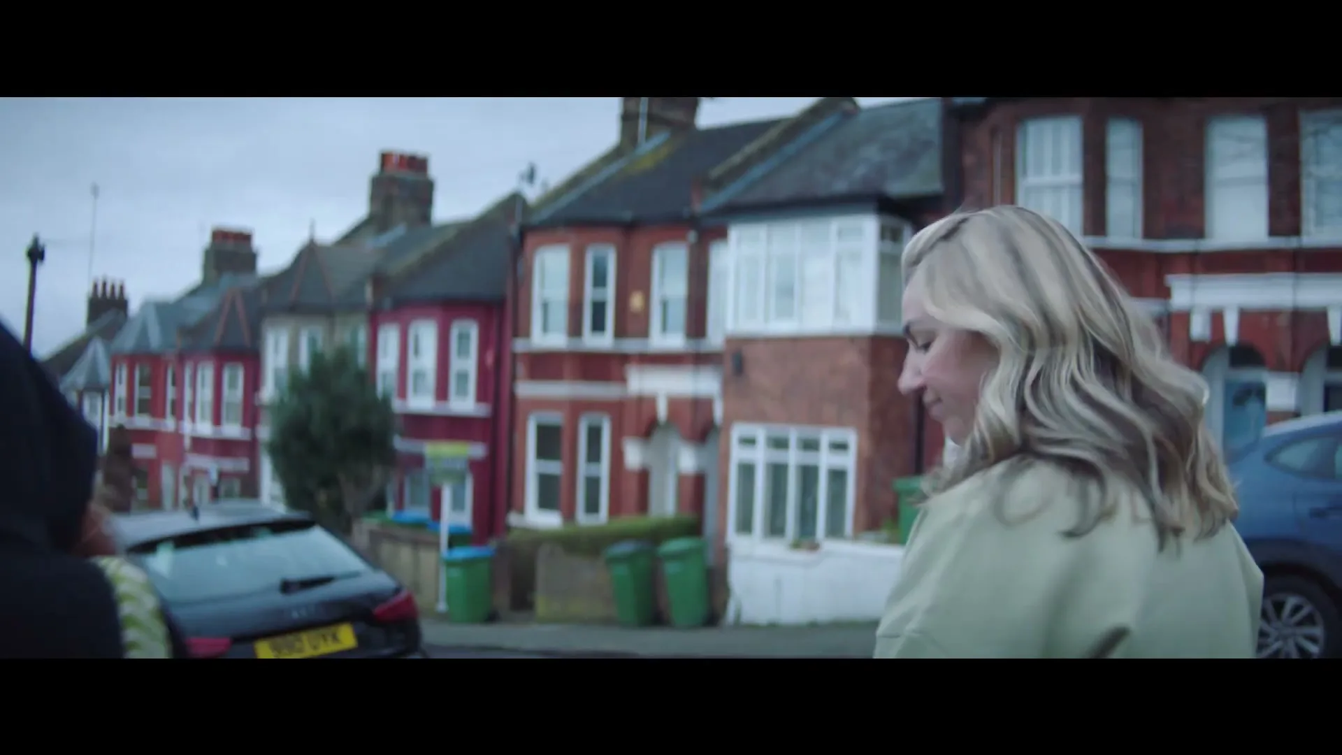 A blonde woman in her 20s walks away from the camera on a residential street with terraced houses in the background.