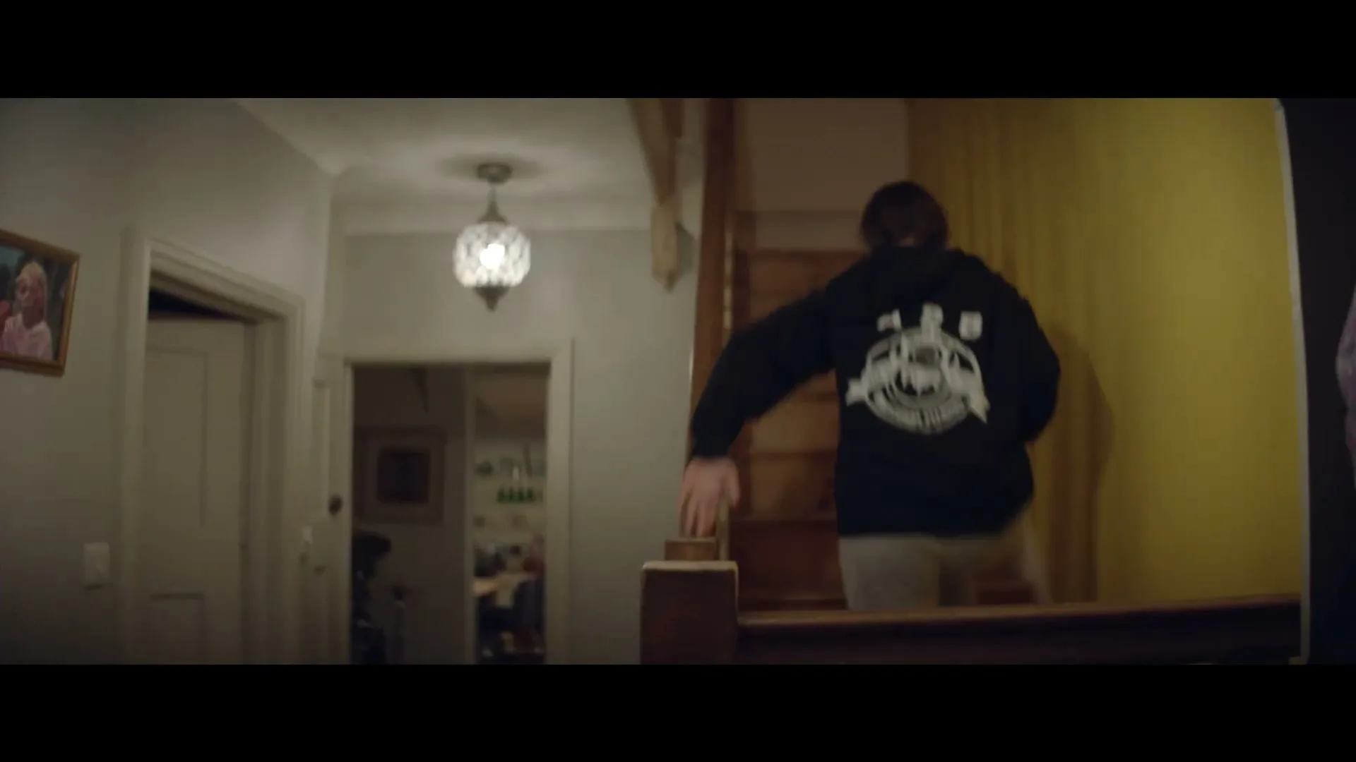 A young person in a black hoodie and grey sweatpants walks up a wooden staircase in a dimly lit house.