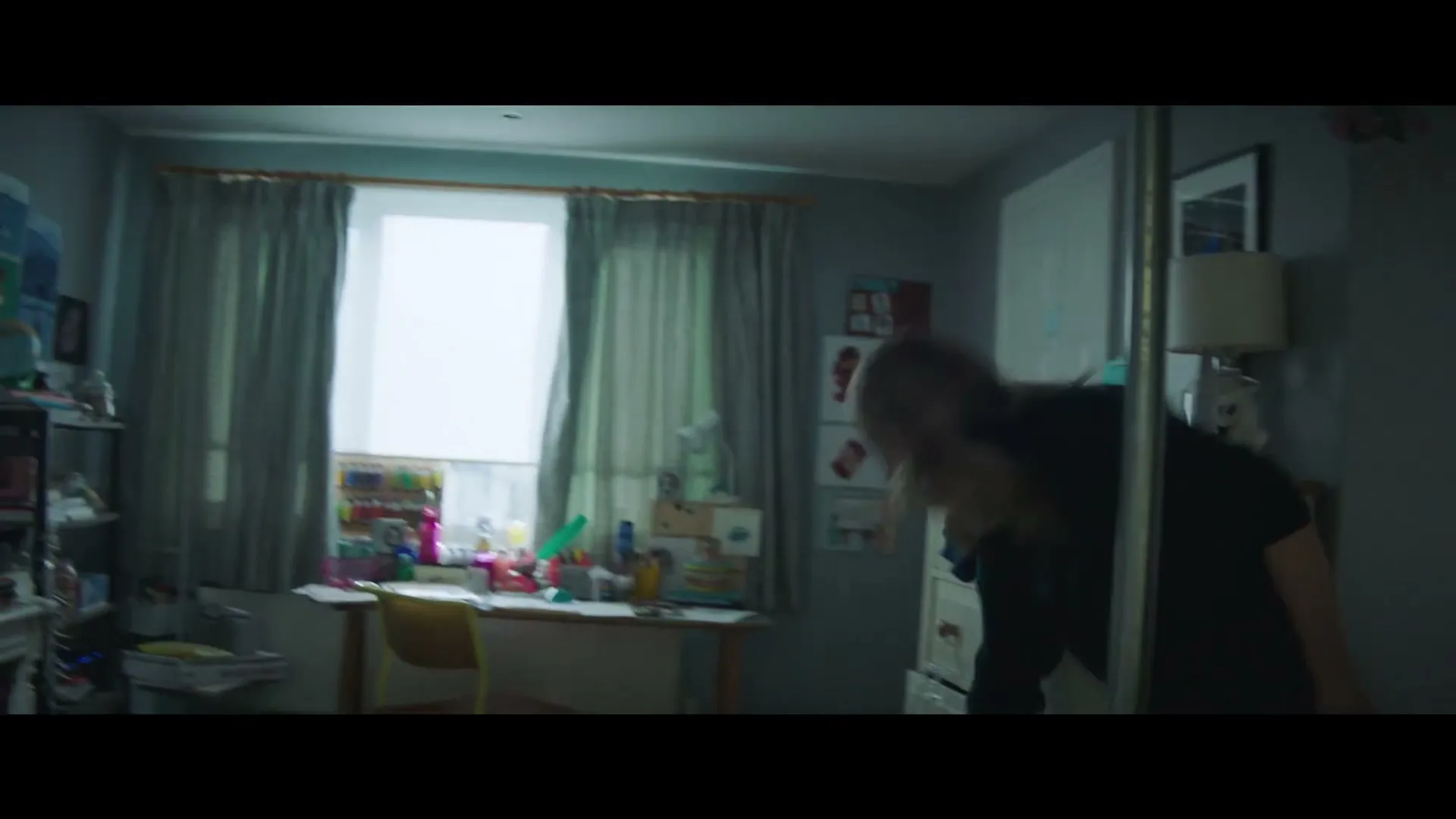 A young woman with blonde hair enters a child's bedroom, bending down to pick something up from the floor.