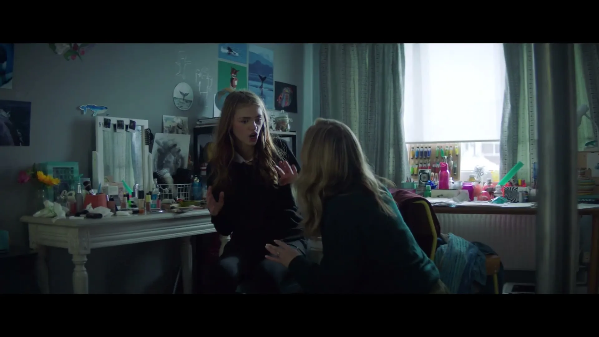 A young woman with long brown hair, wearing a black top, sits on a chair, looking distressed and gesturing with her hands, while another woman kneels beside her, trying to comfort her in a bedroom.