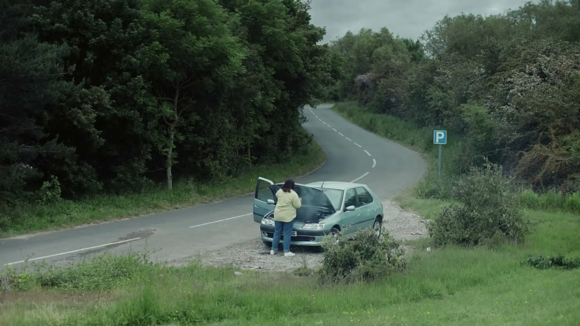 A woman in a yellow jacket stands next to her broken-down car on the side of a winding road, as smoke rises from the engine.