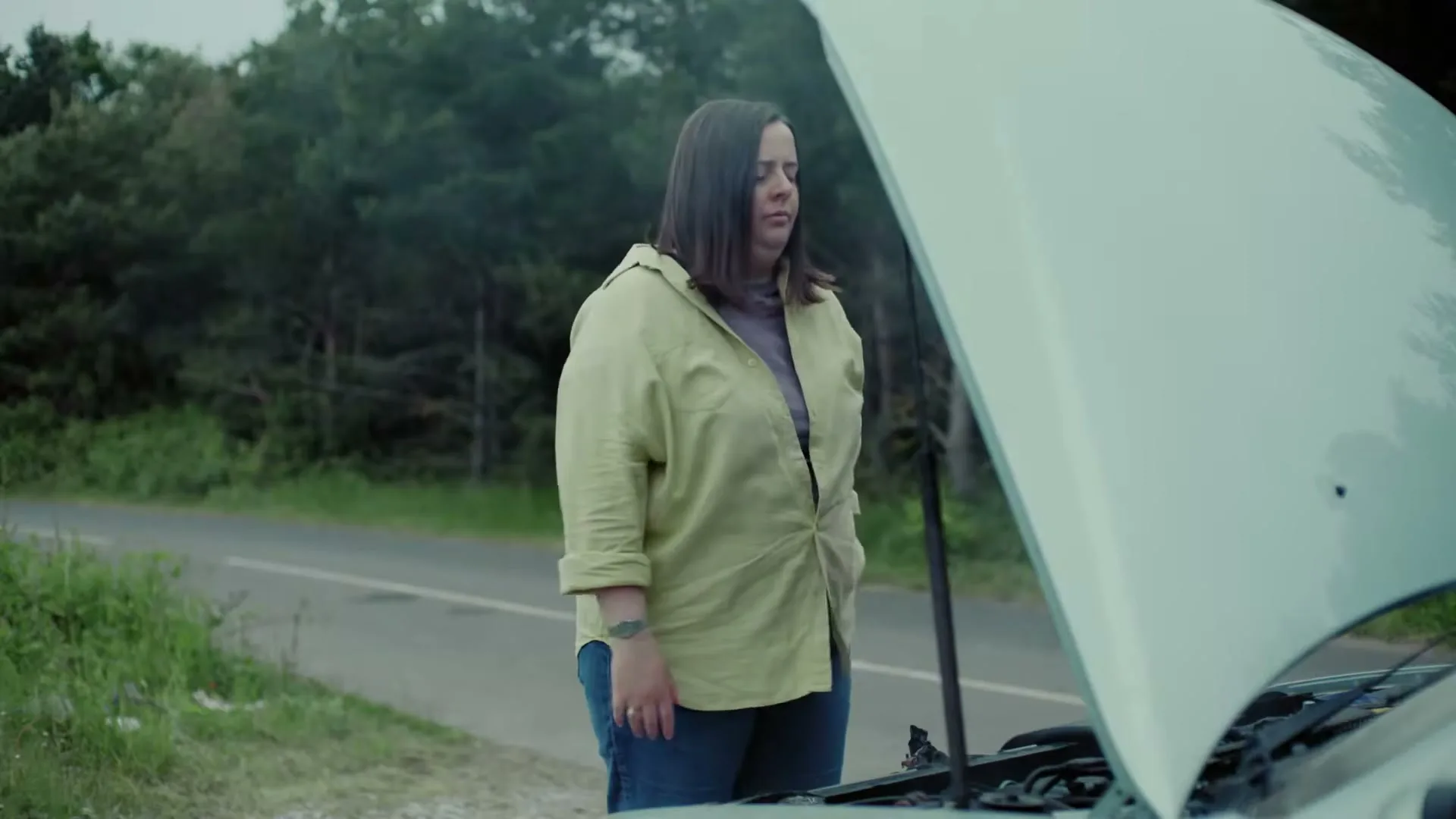 A woman in a yellow shirt and jeans stands next to her broken-down car on the side of a road, looking frustrated as smoke rises from the engine.