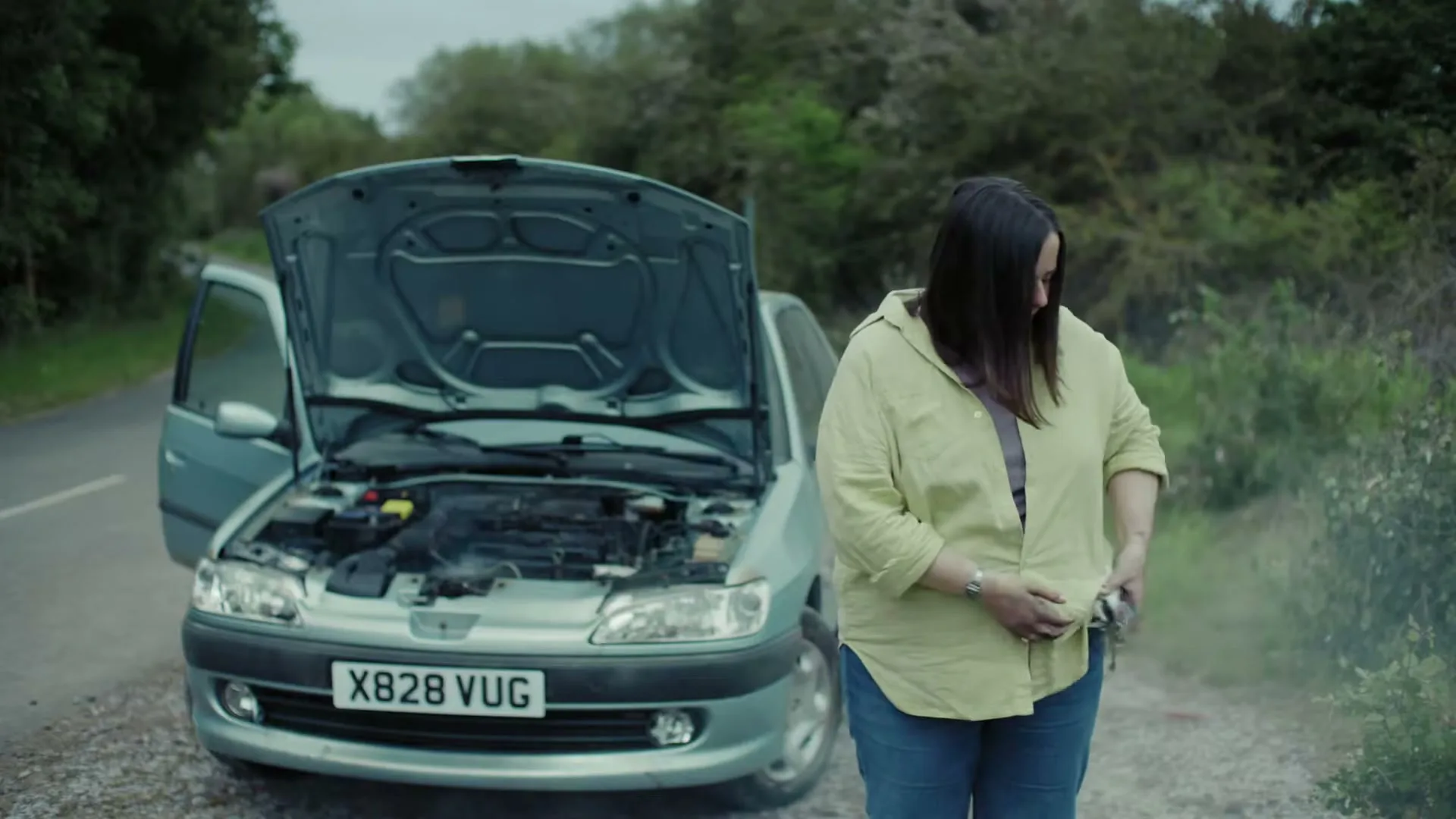 A woman stands next to her broken-down car on a rural road, looking frustrated as she tries to clean her hands with a small cloth.