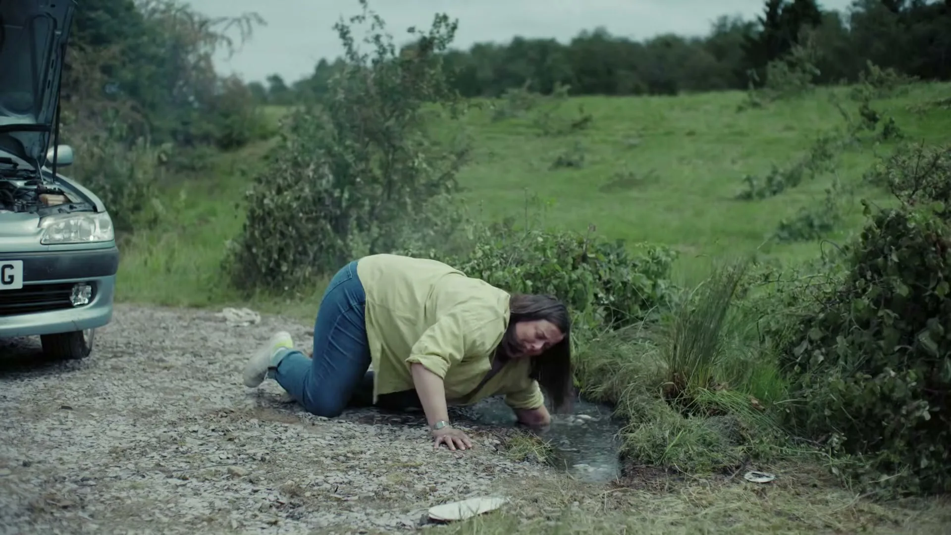 A woman in a yellow shirt and blue jeans is on her hands and knees, looking distressed, next to a broken-down car on a dirt road.