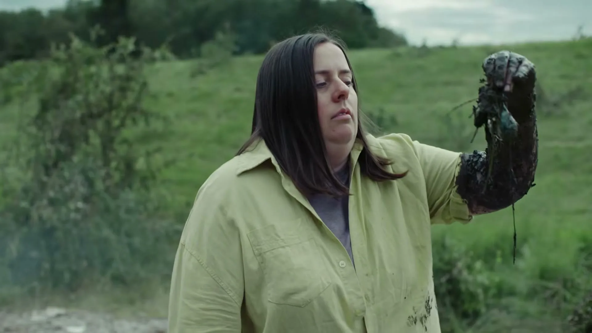 A woman with dark hair and a light green shirt looks at her arm, which is covered in black mud and debris, in a grassy outdoor area.