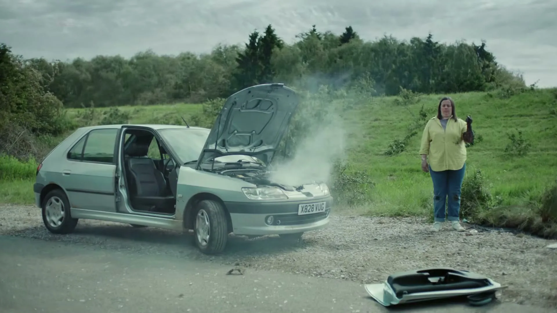 A woman stands next to her broken-down car with smoke coming from the engine, looking frustrated on a rural road.