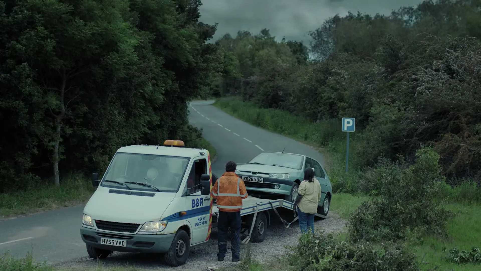 A tow truck driver and a woman stand next to a broken-down car being loaded onto a tow truck on a rural road.