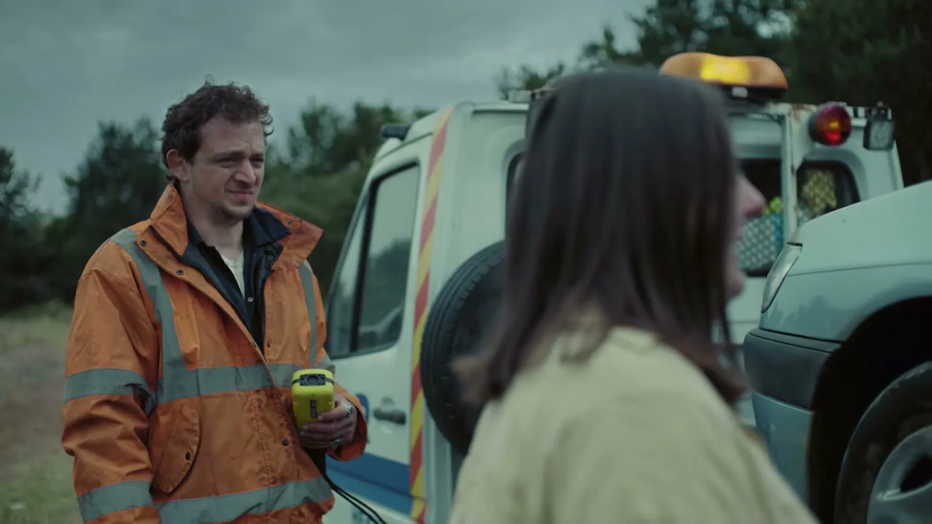 A male tow truck driver in an orange jacket holds a device, looking at a woman whose back is to the camera, next to a broken-down car on a dirt road.