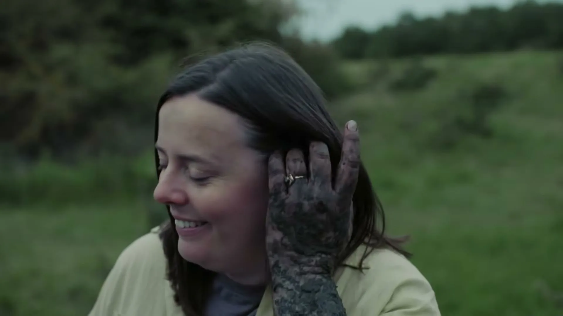 A woman with dark hair and a dirty hand smiles and puts an earbud in her ear while standing in a grassy field.