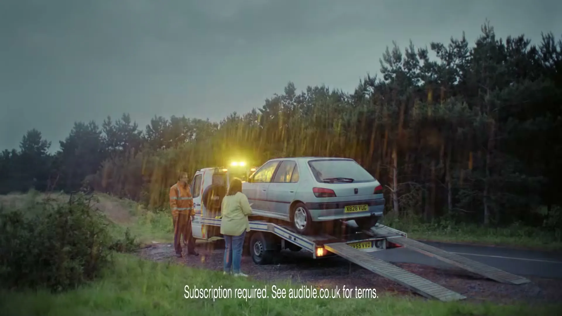 A tow truck driver and a woman stand next to a broken-down car on a tow truck, on a rural road as lightning strikes.