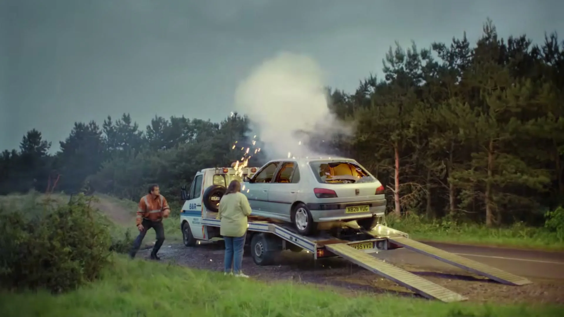 A tow truck driver and a woman watch as a car on a tow truck bursts into flames on a rural road at dusk.