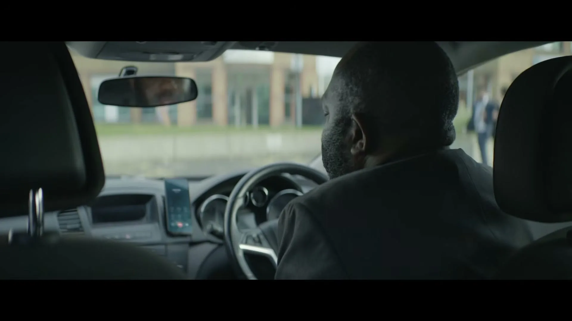 A man with dark skin and a beard, wearing a suit, sits in the driver's seat of a car, looking over his shoulder as he talks on the phone.