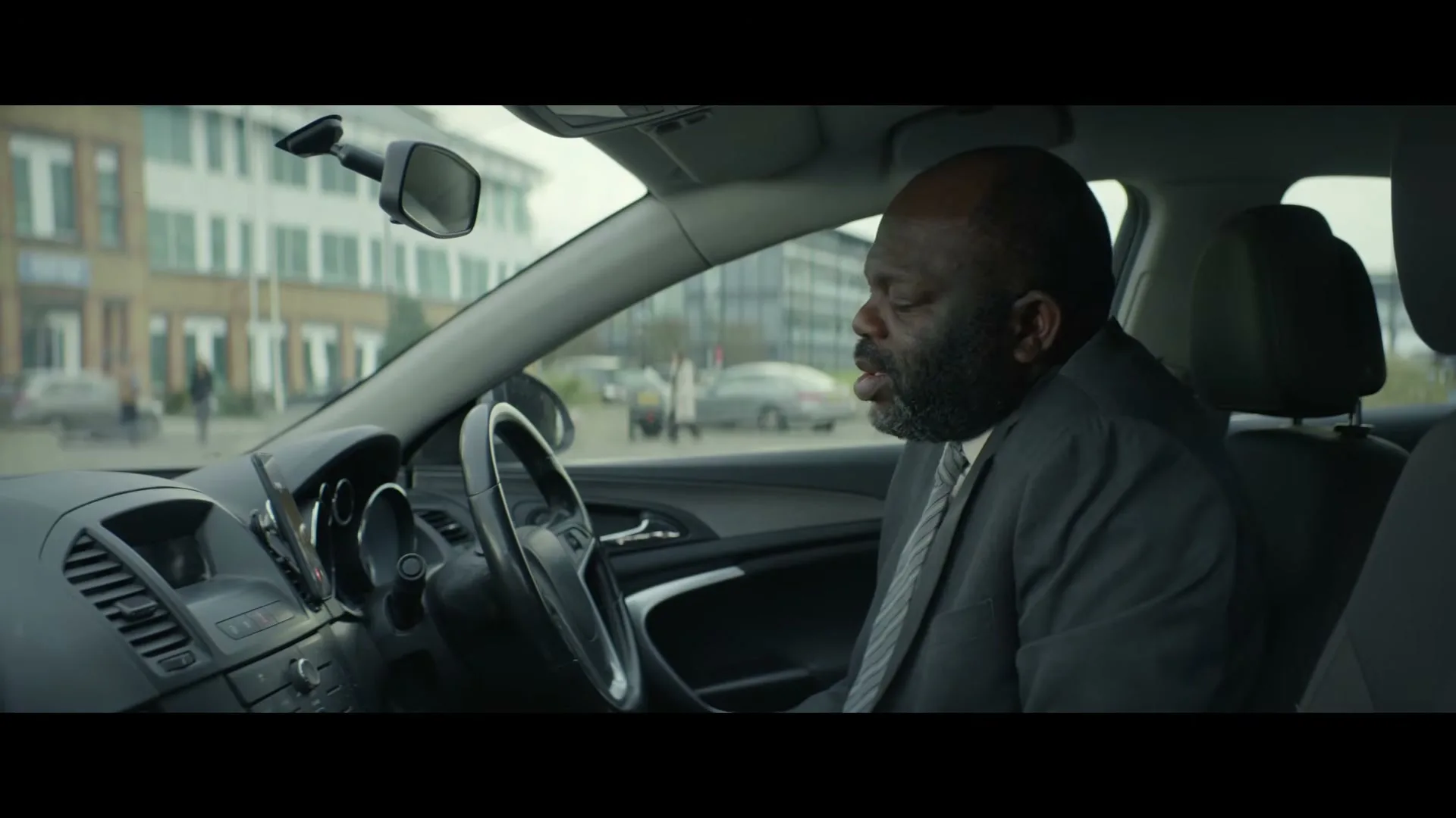 A middle-aged Black man in a suit sits in the driver's seat of a car, looking down and talking on speakerphone in a parking lot.