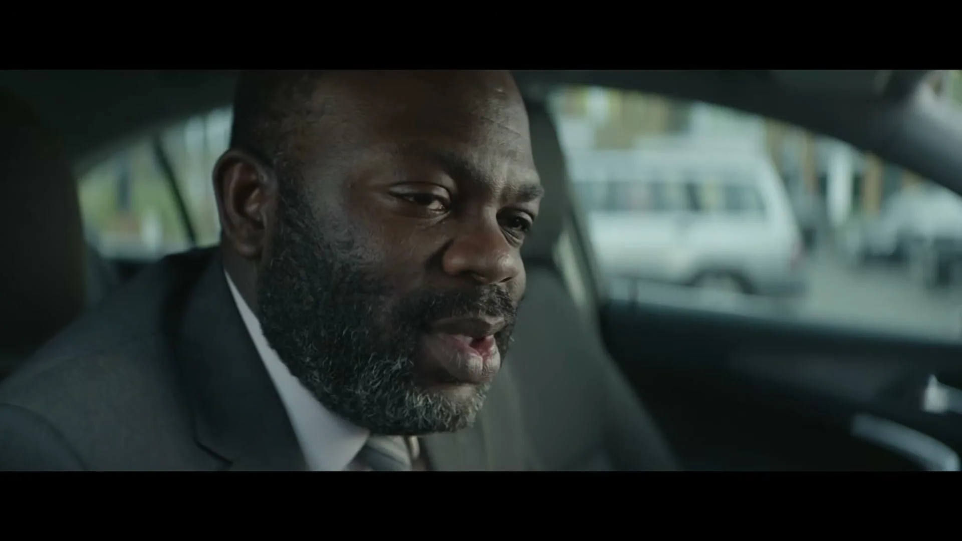 A middle-aged Black man with a beard, wearing a suit, sits in a car and looks down, then turns to his right.
