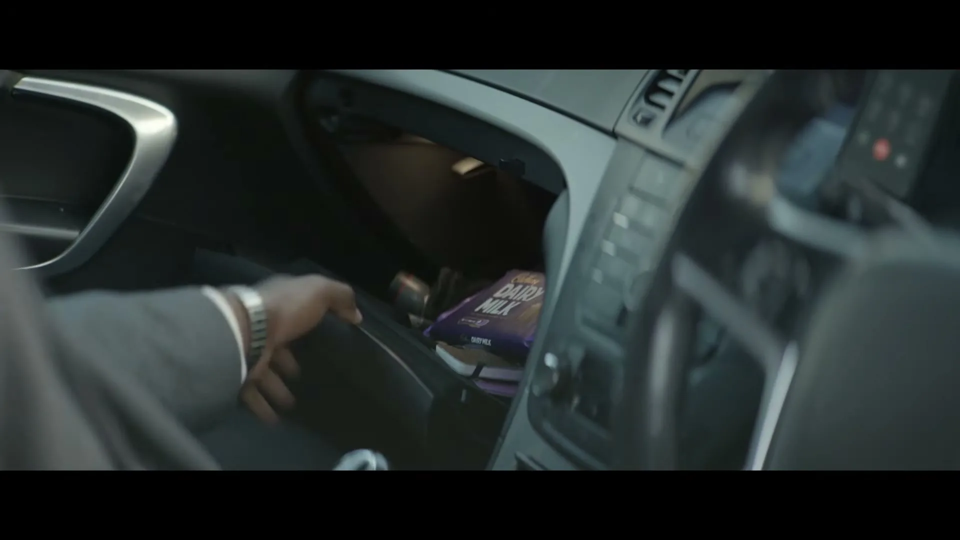 A man in a suit opens the glove compartment of his car and pulls out a purple Cadbury Dairy Milk chocolate bar.