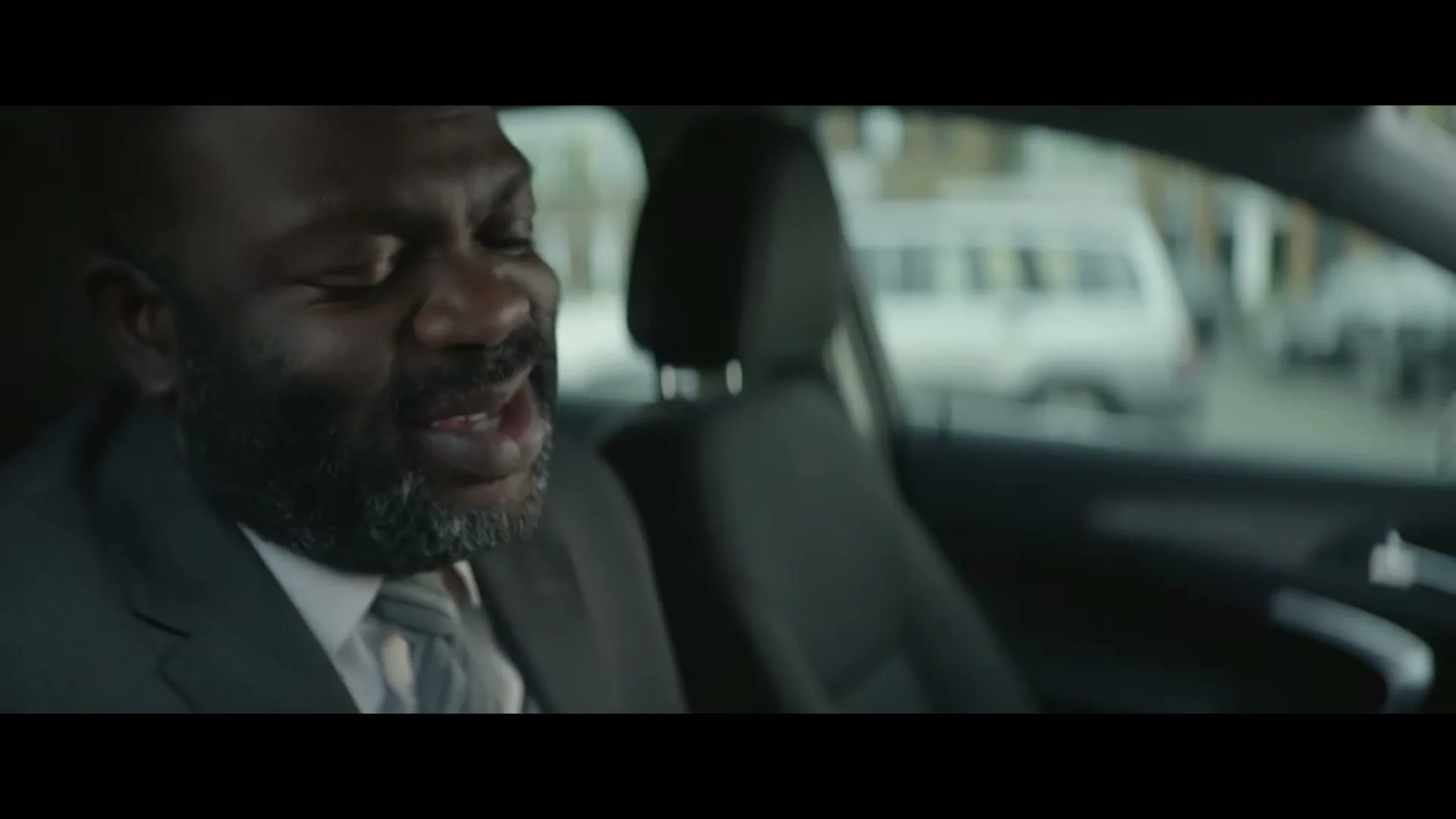 A Black man in his 40s, wearing a suit and tie, sits in the driver's seat of a car, looking down and talking.