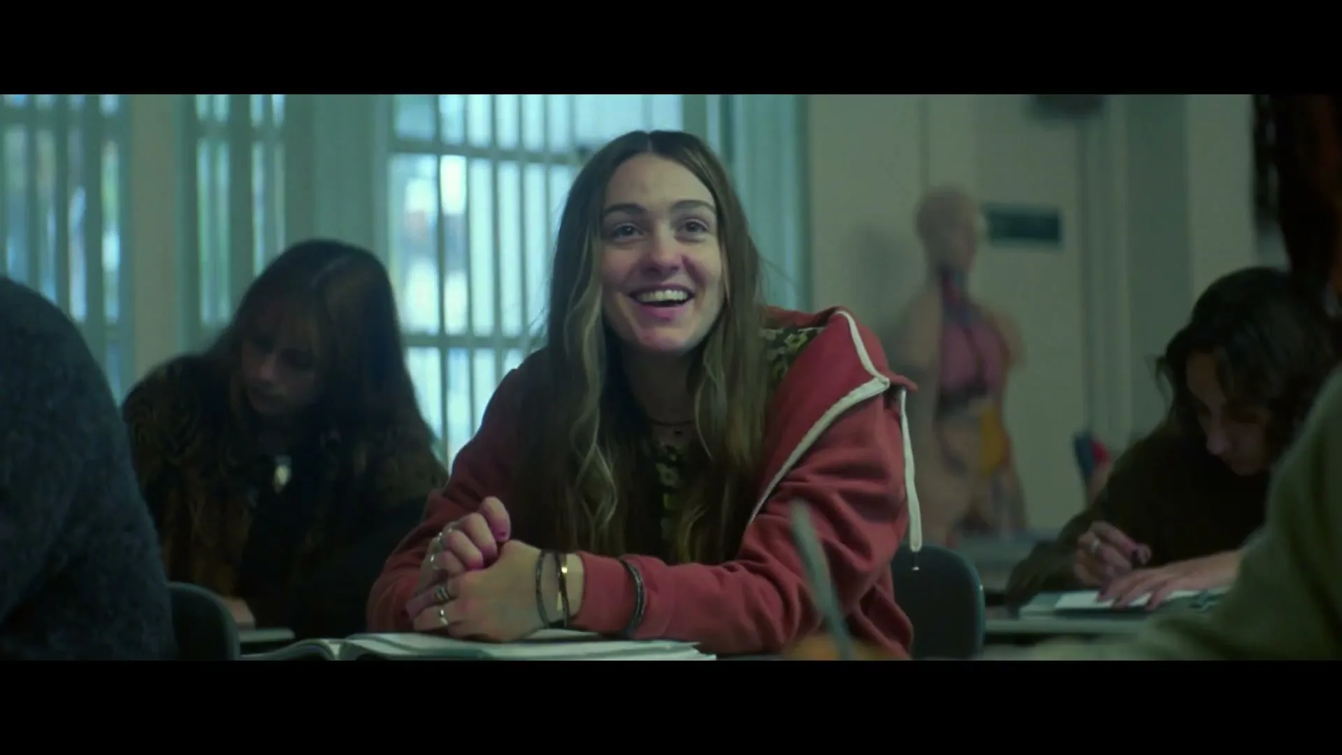 A young woman with long brown hair, wearing a red hoodie, smiles brightly while sitting at a desk in a classroom with other students.