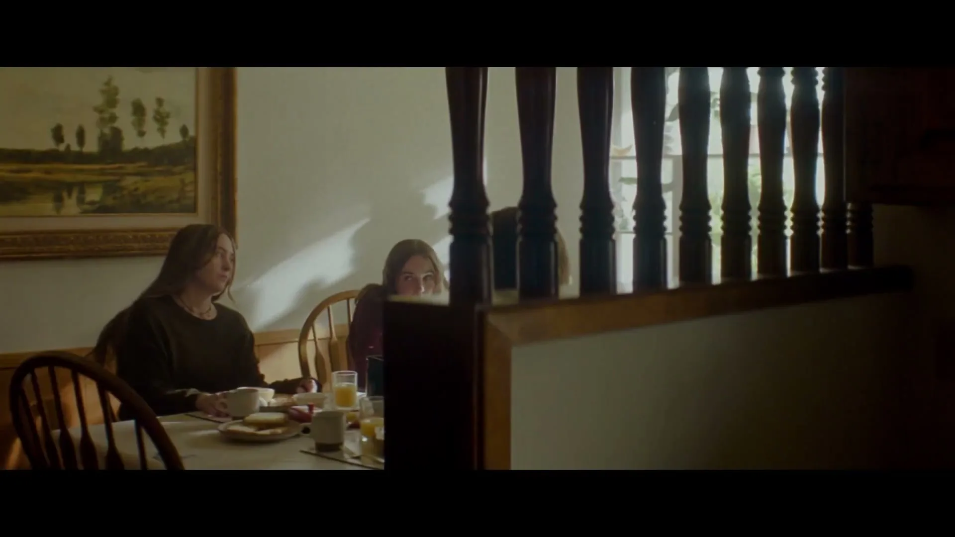 Two young women sit at a dining table, partially obscured by a wooden banister, having breakfast in a dimly lit room.