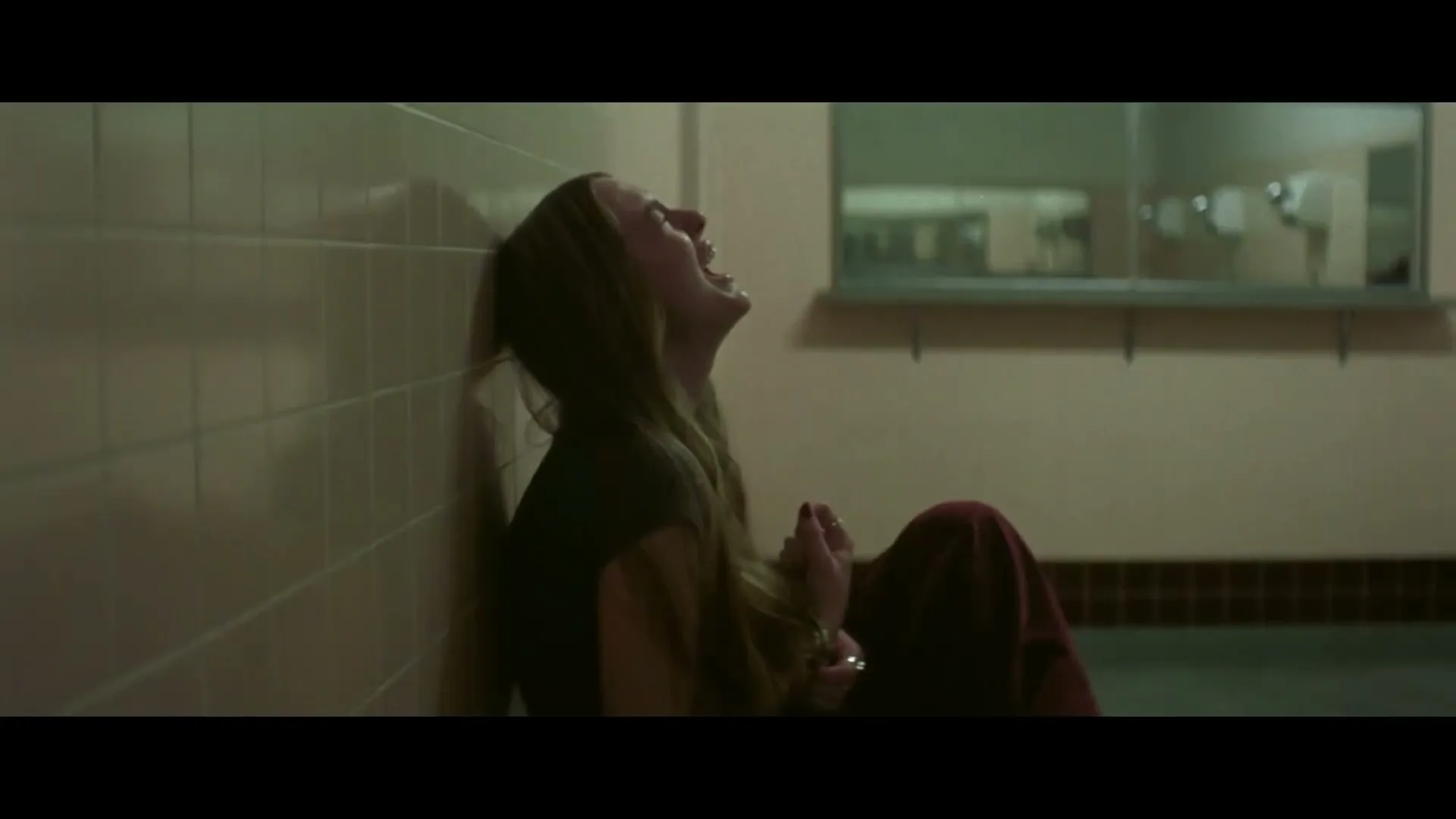 A young woman with long brown hair, wearing a dark top and red pants, sits on the floor crying in a dimly lit hallway.