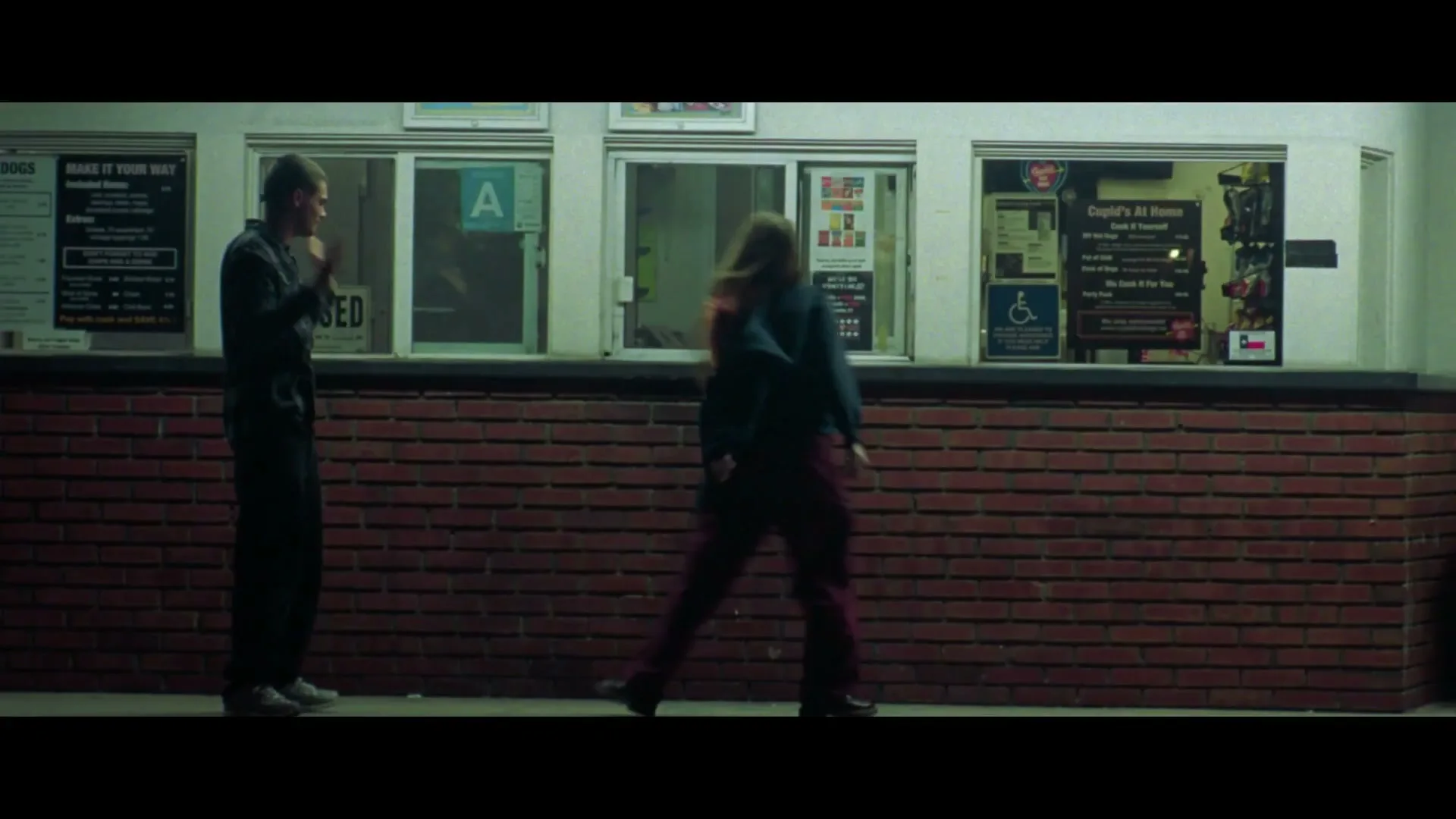 A young man stands outside a fast-food restaurant at night, looking down as a young woman walks past him, her face obscured.